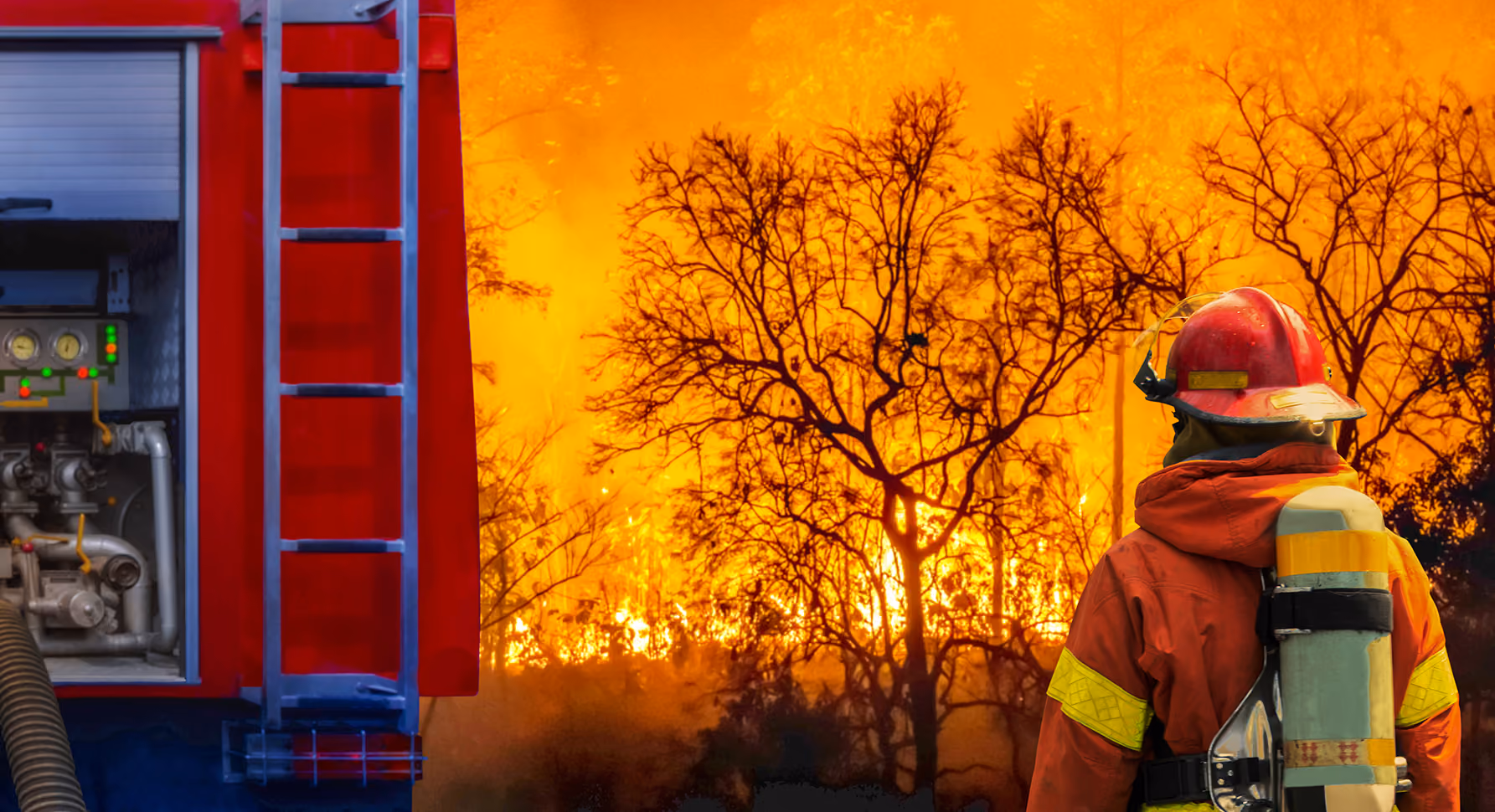 Firefighter faces an intense wildfire burning through trees beside a fire engine.