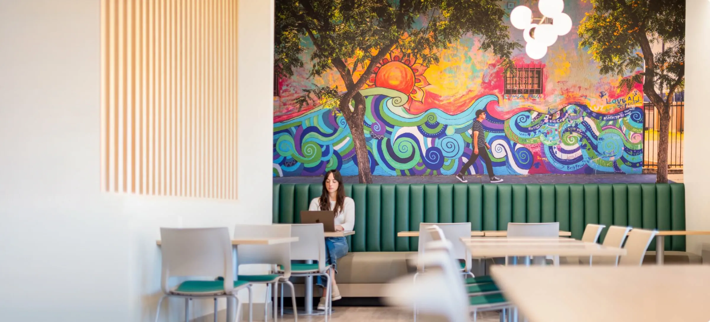 Woman sits beneath a large colorful mural in a bright community gathering space.