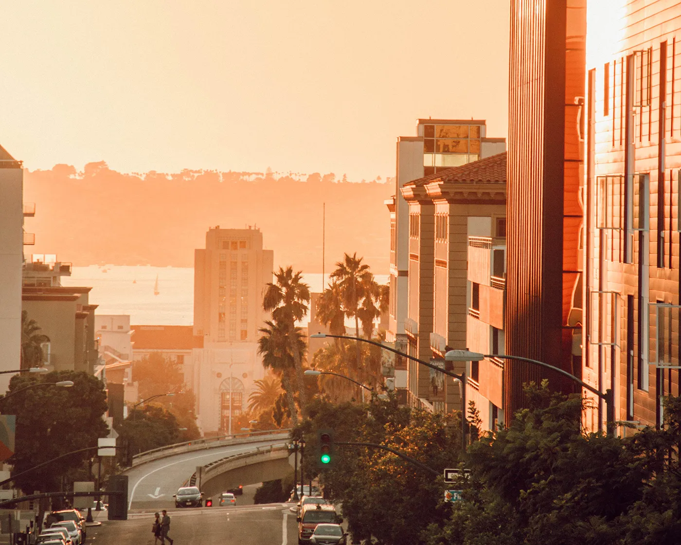 Sunlit city street slopes toward the distant coastline, with buildings and palm trees.