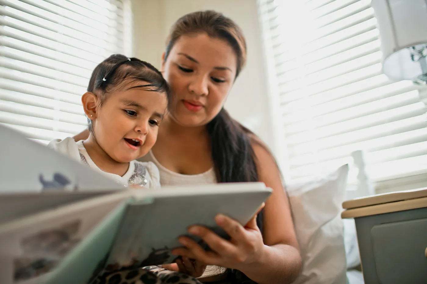 Mother and young daughter smile while reading a book together at home.