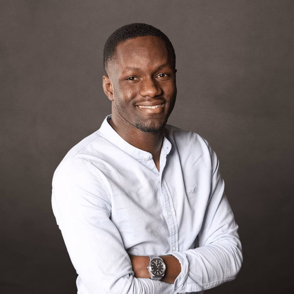 Portrait of a smiling man with short hair wearing a white buttoned shirt and a wristwatch, standing with arms crossed against a dark neutral background.