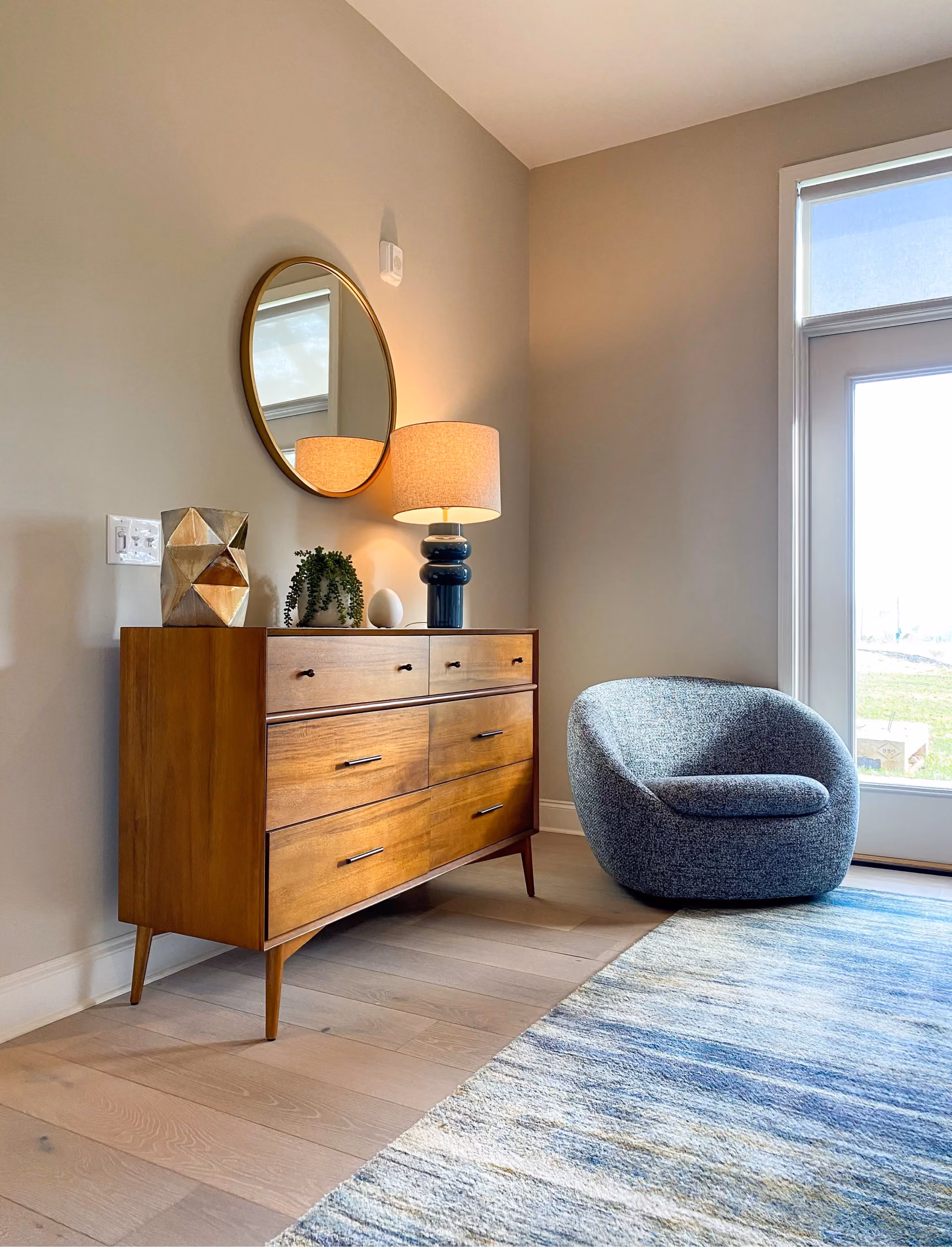 Student apartment bedroom with a wood dresser, round mirror, and blue accent chair