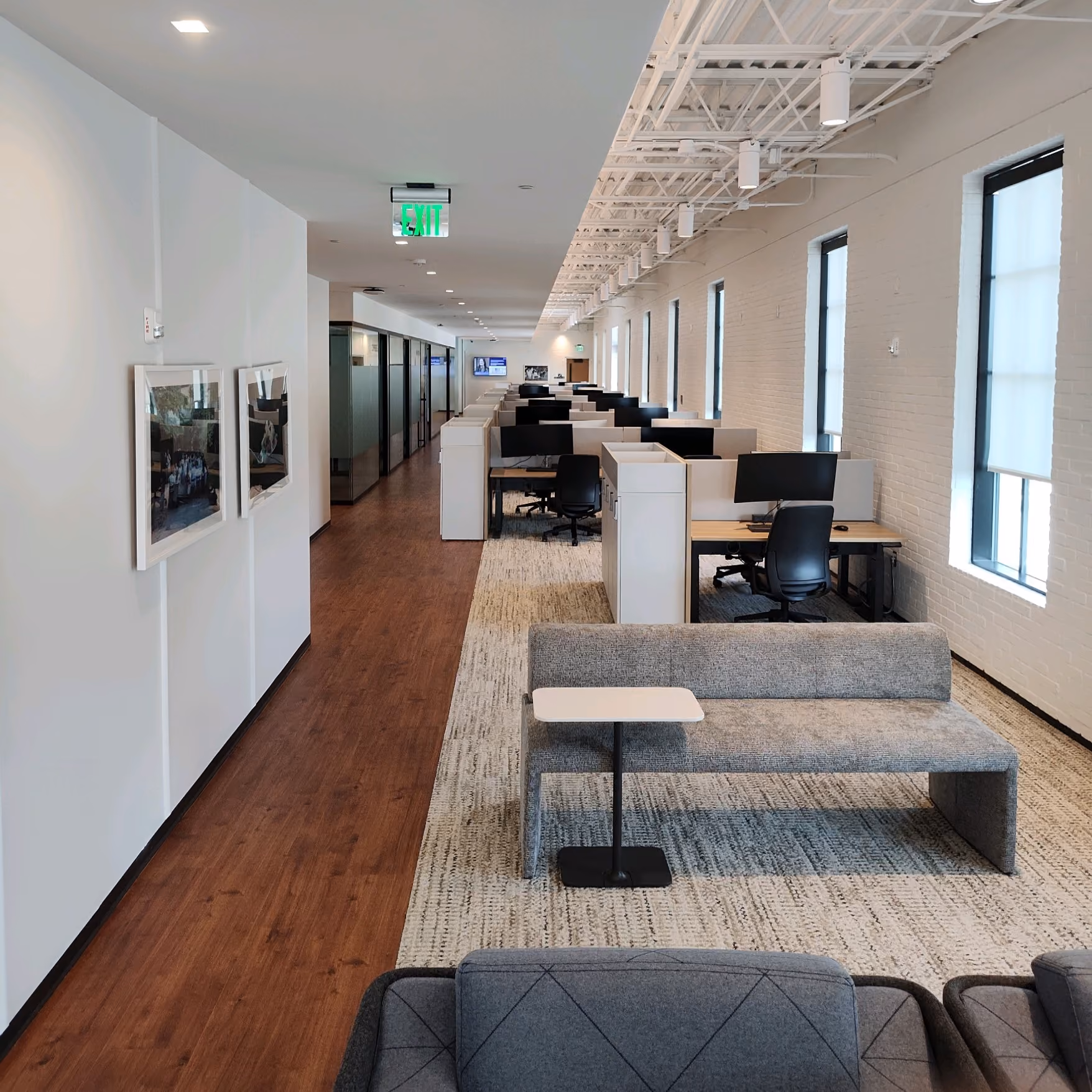 Office hallway with workstations along a wall on dark wood-look flooring and framed wall art