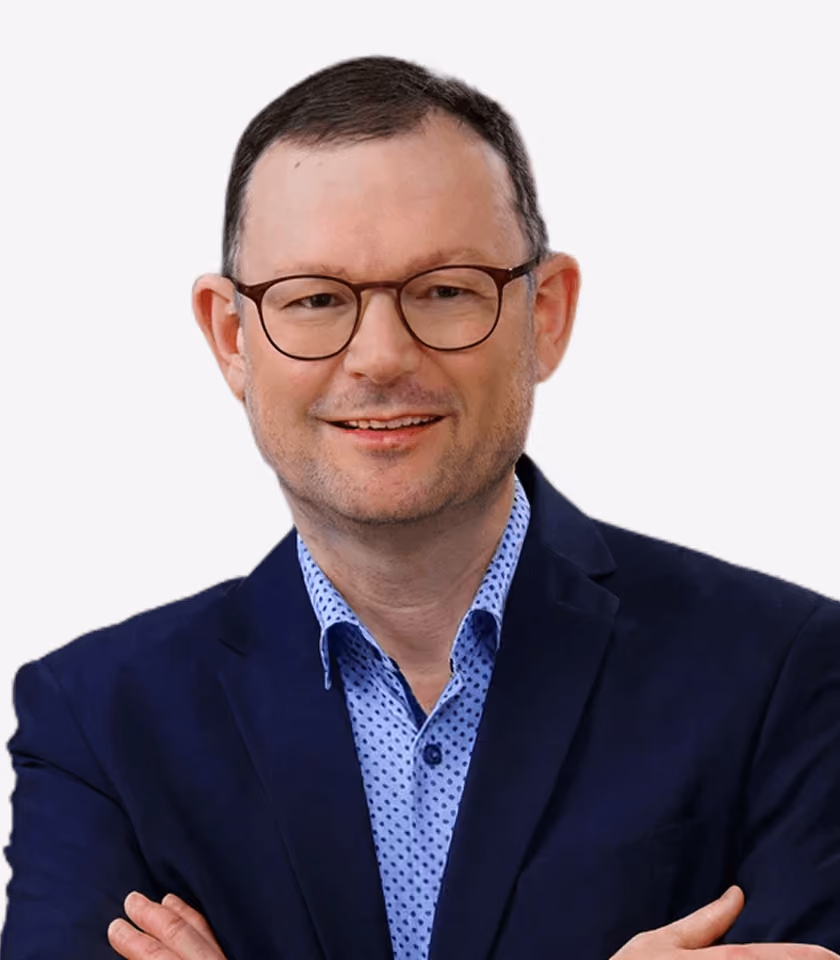 Mann mit Brille und blauem Anzug lächelt und verschränkt die Arme vor weißem Hintergrund.