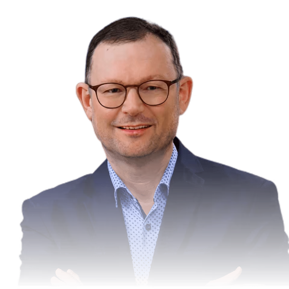 Mann mit Brille, im blauen Anzug, lächelnd mit verschränkten Armen vor weißem Hintergrund.