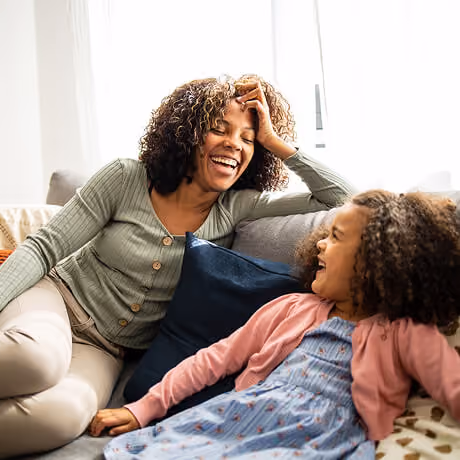 A smiling woman with curly hair wearing a green buttoned top sits on a couch, leaning towards a young girl with curly hair in a blue dress and pink cardigan, both sharing a joyful moment.