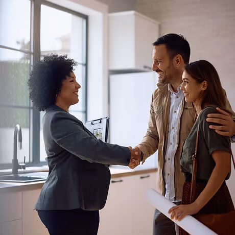 A real estate agent with curly hair shaking hands with a smiling couple in a modern kitchen, while the woman holds architectural plans.