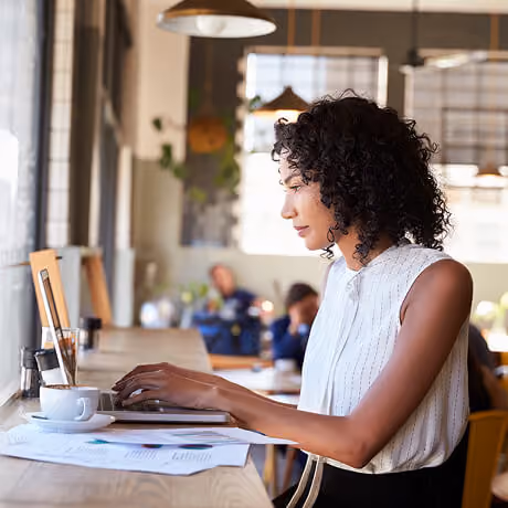 A woman with curly hair in a sleeveless white top is sitting at a wooden counter typing on a laptop, with papers and a cup of coffee beside her, in a bright café setting.
