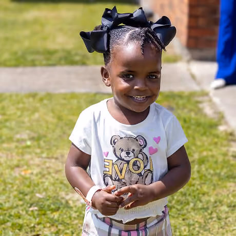 Smiling young girl standing outdoors on grass wearing a white shirt with a teddy bear and the word LOVE, with large black bows in her braided hair.