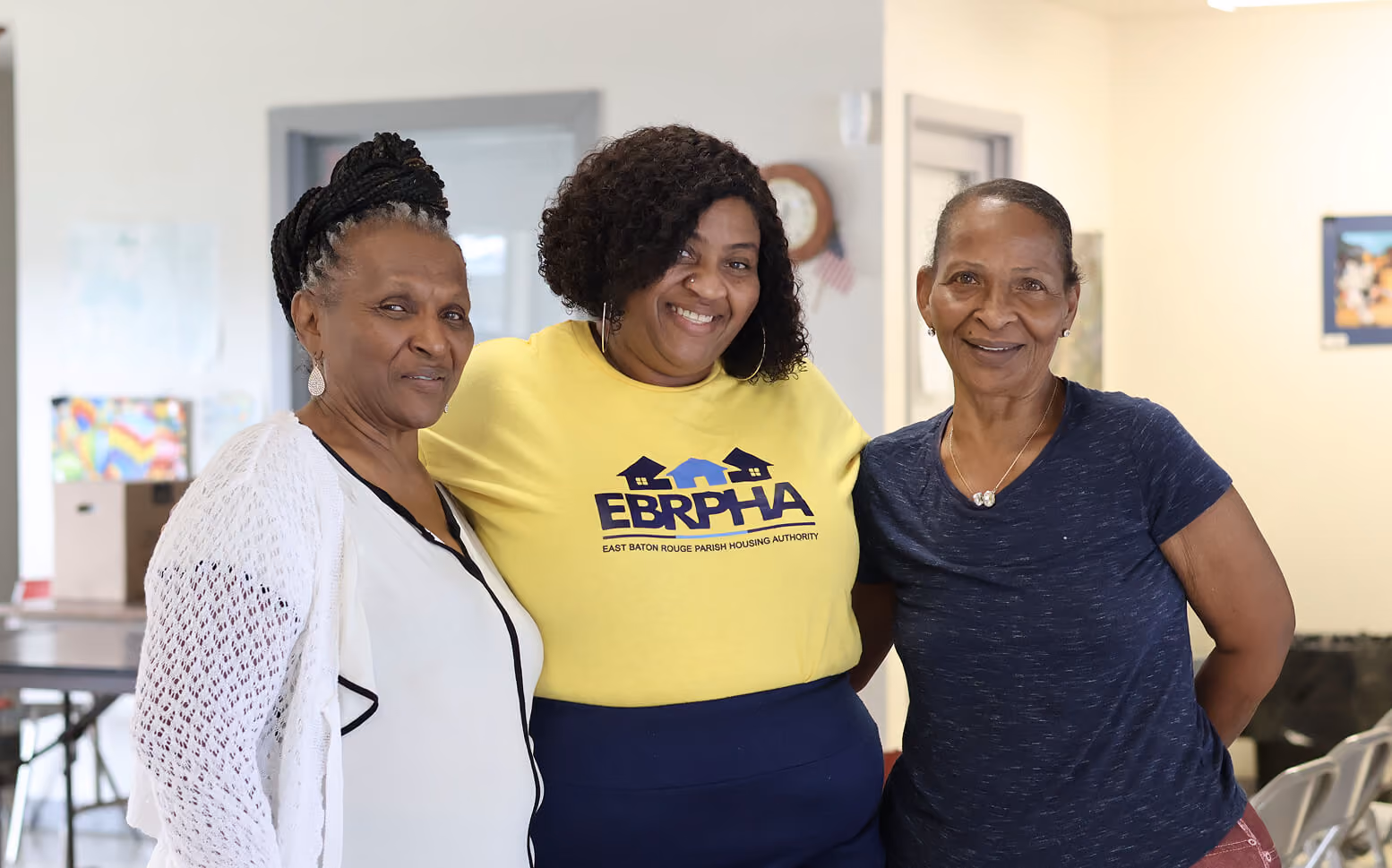 Three smiling women standing together indoors posing for the photo; the woman in the middle wears a yellow shirt with the East Baton Rouge Parish Housing Authority logo.