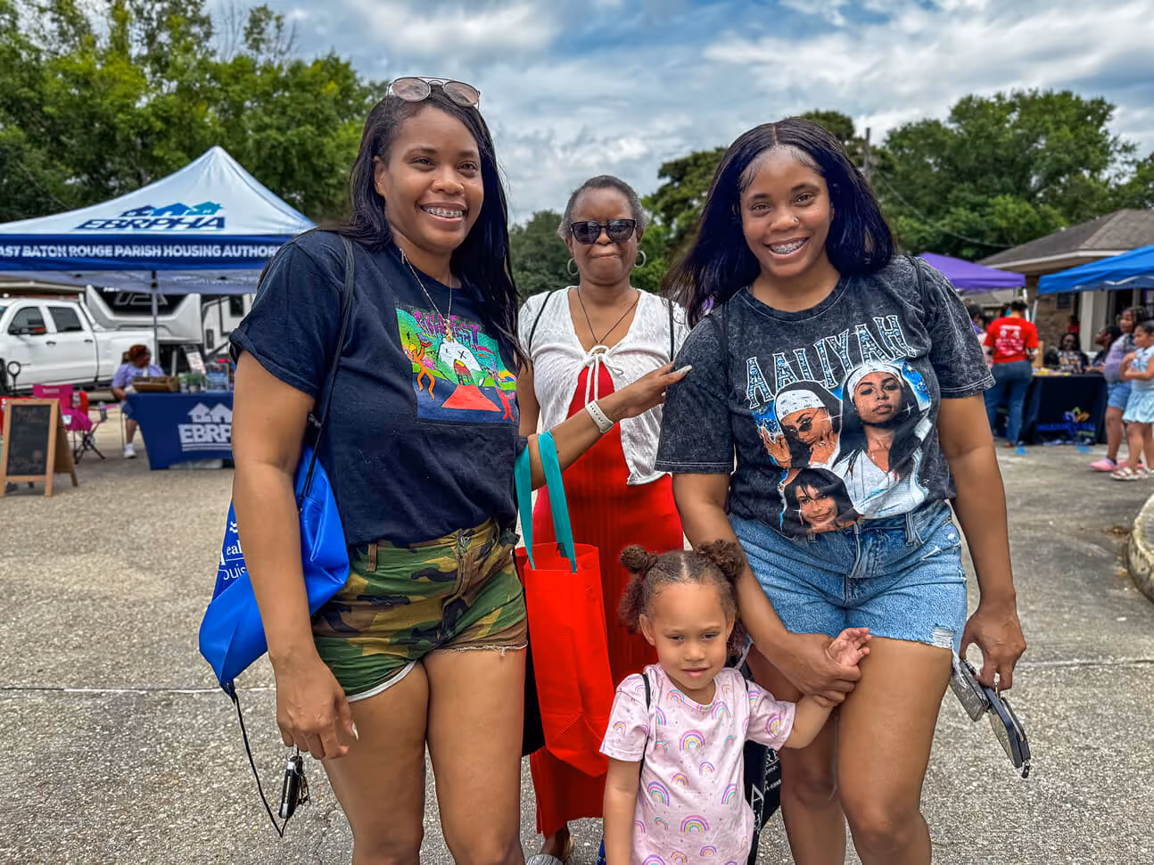 Four Black females smiling outdoors at a community event, including a young girl with rainbow shirt holding hands with a woman in an Aaliyah graphic t-shirt and denim shorts, another woman in a black graphic t-shirt and camo shorts holding a blue bag, and an older woman wearing sunglasses and a red dress in the background near a tent that reads East Baton Rouge Parish Housing Authority.