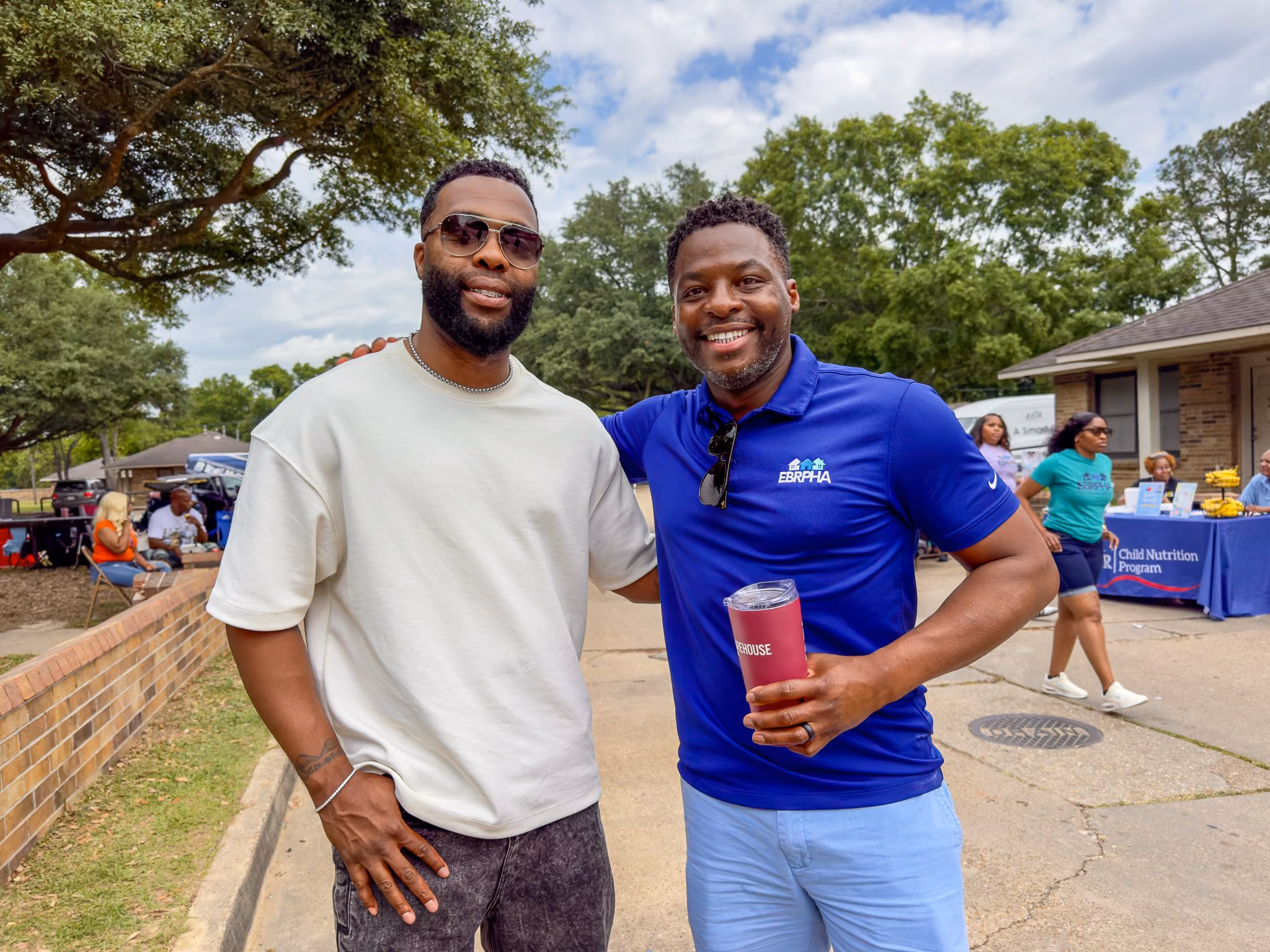 Two men smiling outdoors; the man on the left wears sunglasses and a white T-shirt, the man on the right wears a blue EBRPHA polo shirt holding a red tumbler. In the background, several people are near a table with a Child Nutrition Program banner.