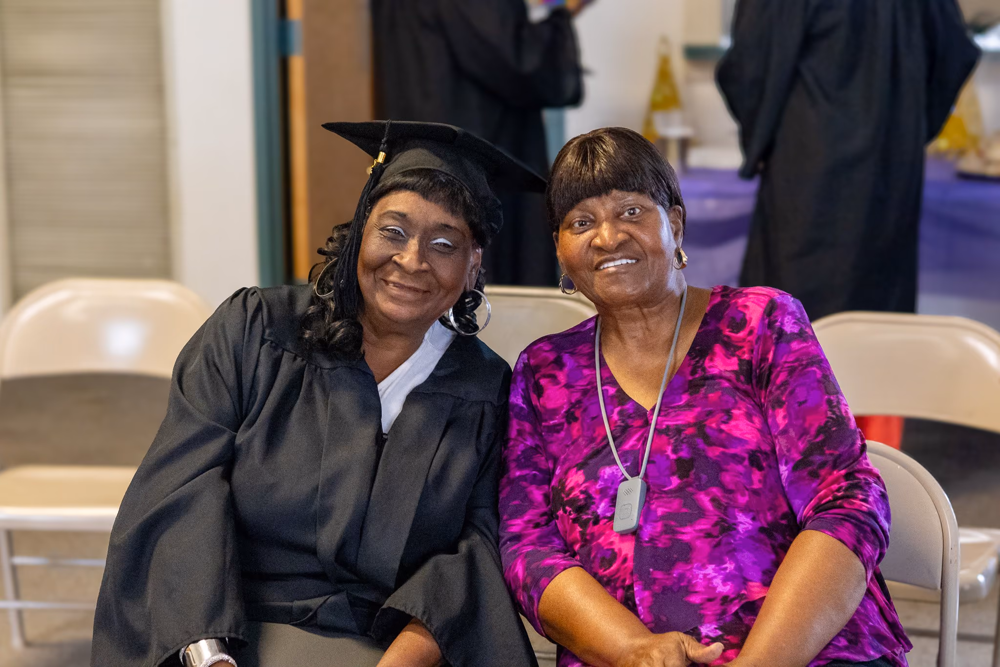 Two smiling older African American women sitting next to each other on beige chairs, one wearing a black graduation gown and cap, the other in a purple patterned blouse with a necklace device.