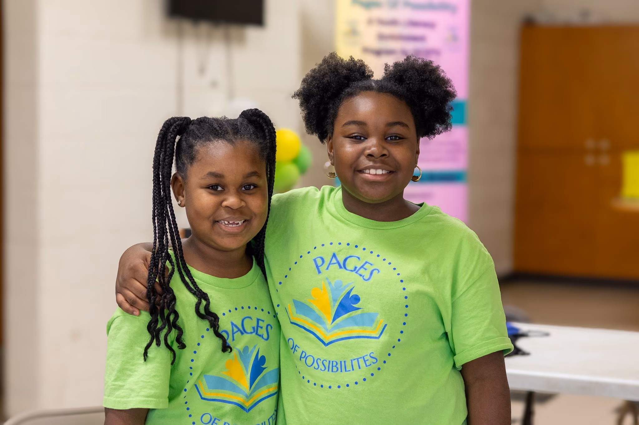 Two smiling young girls with braided hair and afro puffs wearing matching bright green t-shirts that say 'Pages of Possibilities' with a colorful book logo, standing indoors with one arm around the other's shoulder.
