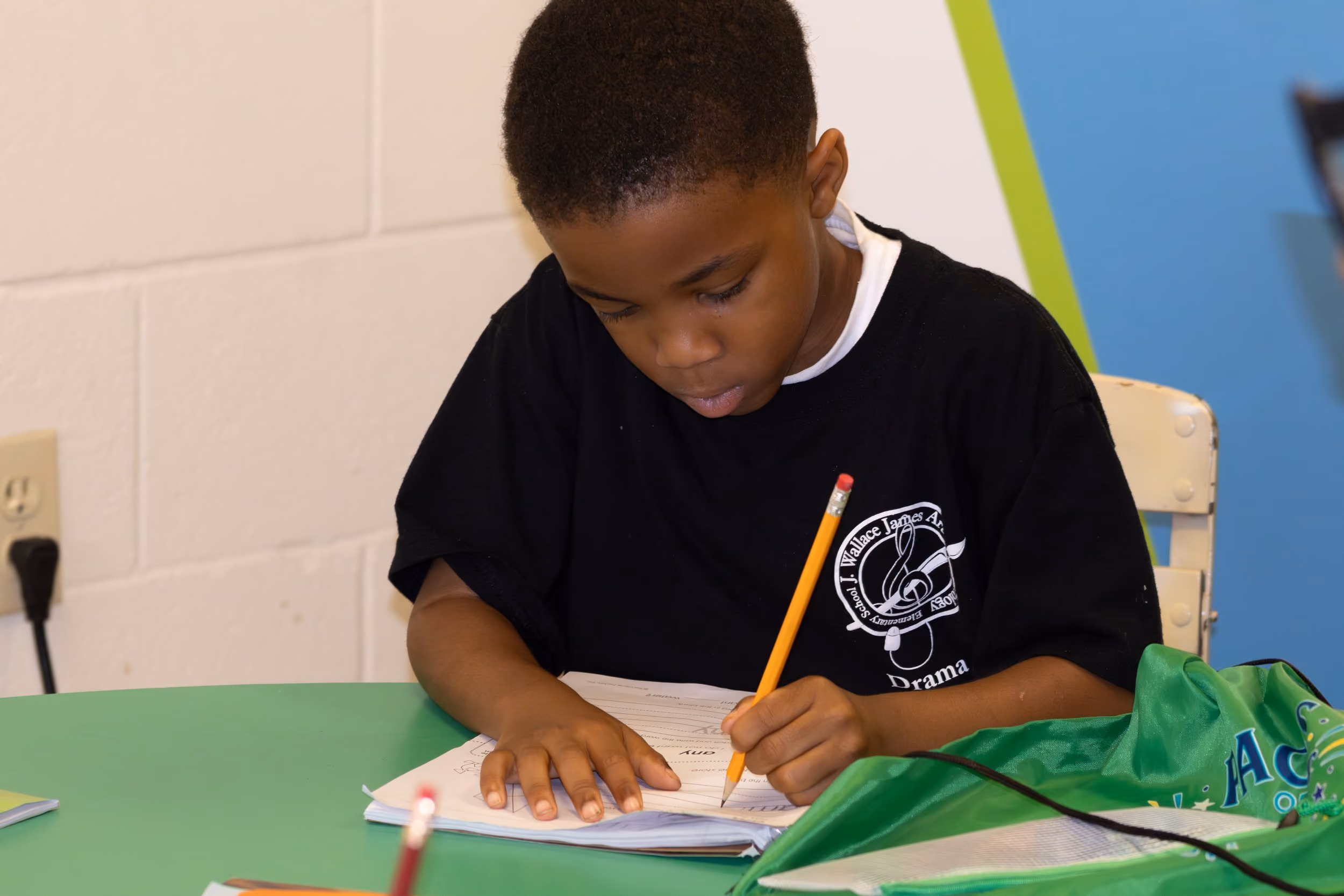 A boy with short hair wearing a black T-shirt is sitting at a green table writing in a notebook with a pencil.