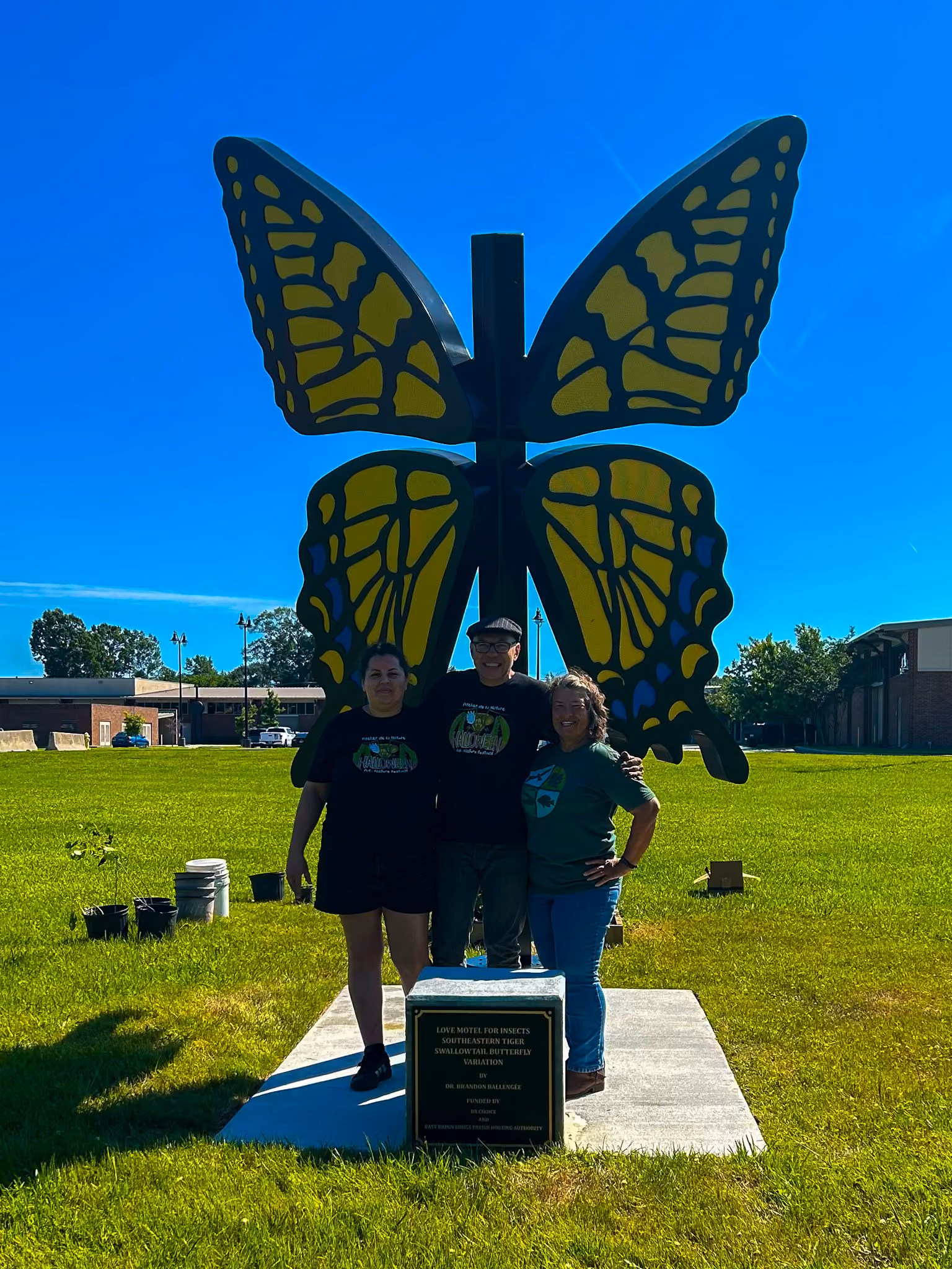 Three people standing in front of a large butterfly sculpture with yellow and black wings on a grassy lawn under a clear blue sky, with a plaque in front mentioning a Love Motel for Insects, Southeastern Tiger Swallowtail Butterfly variation.