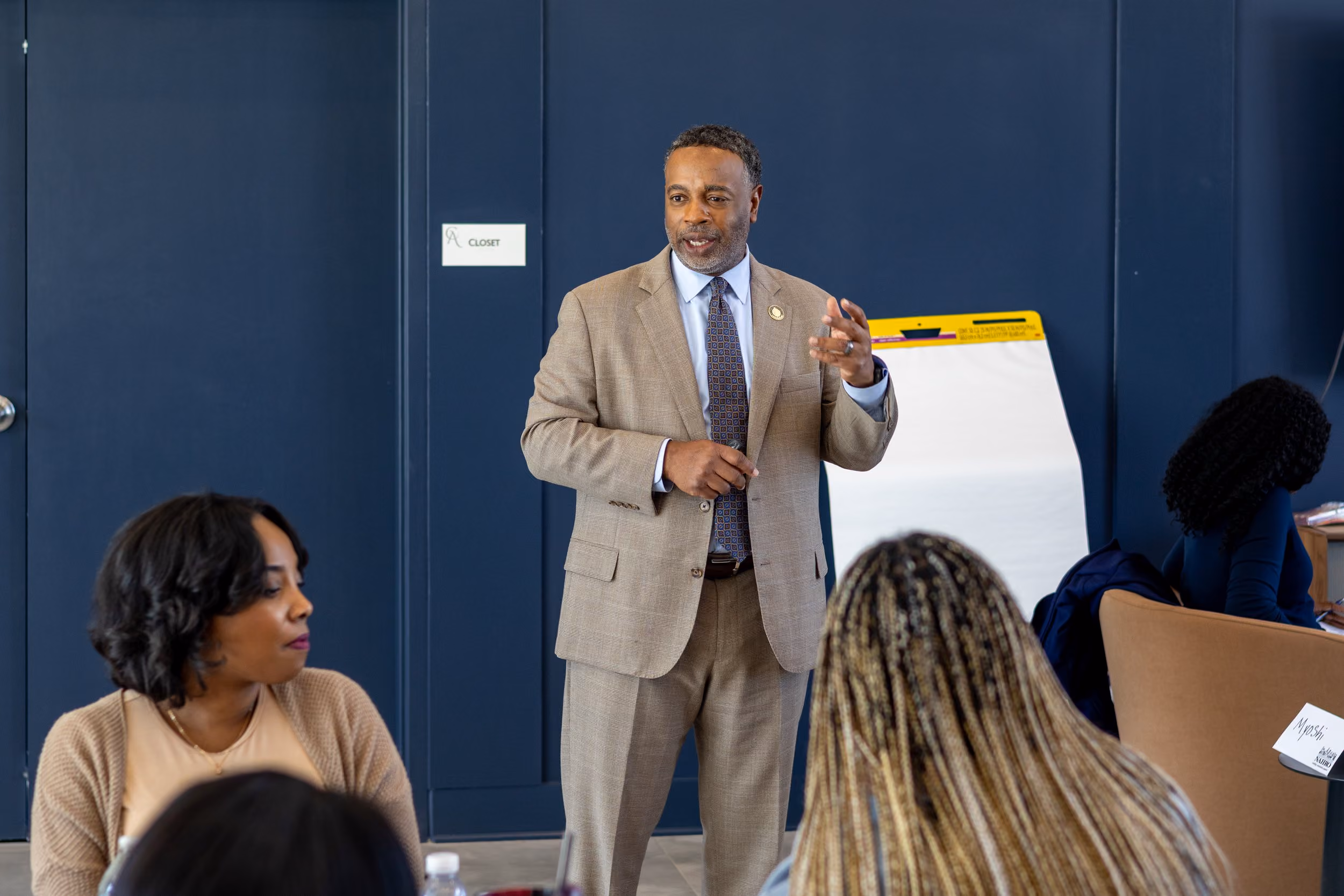 A man in a beige suit and patterned tie is standing and speaking to a group of people seated around him in a room with dark blue walls and a blank flip chart stand behind him.