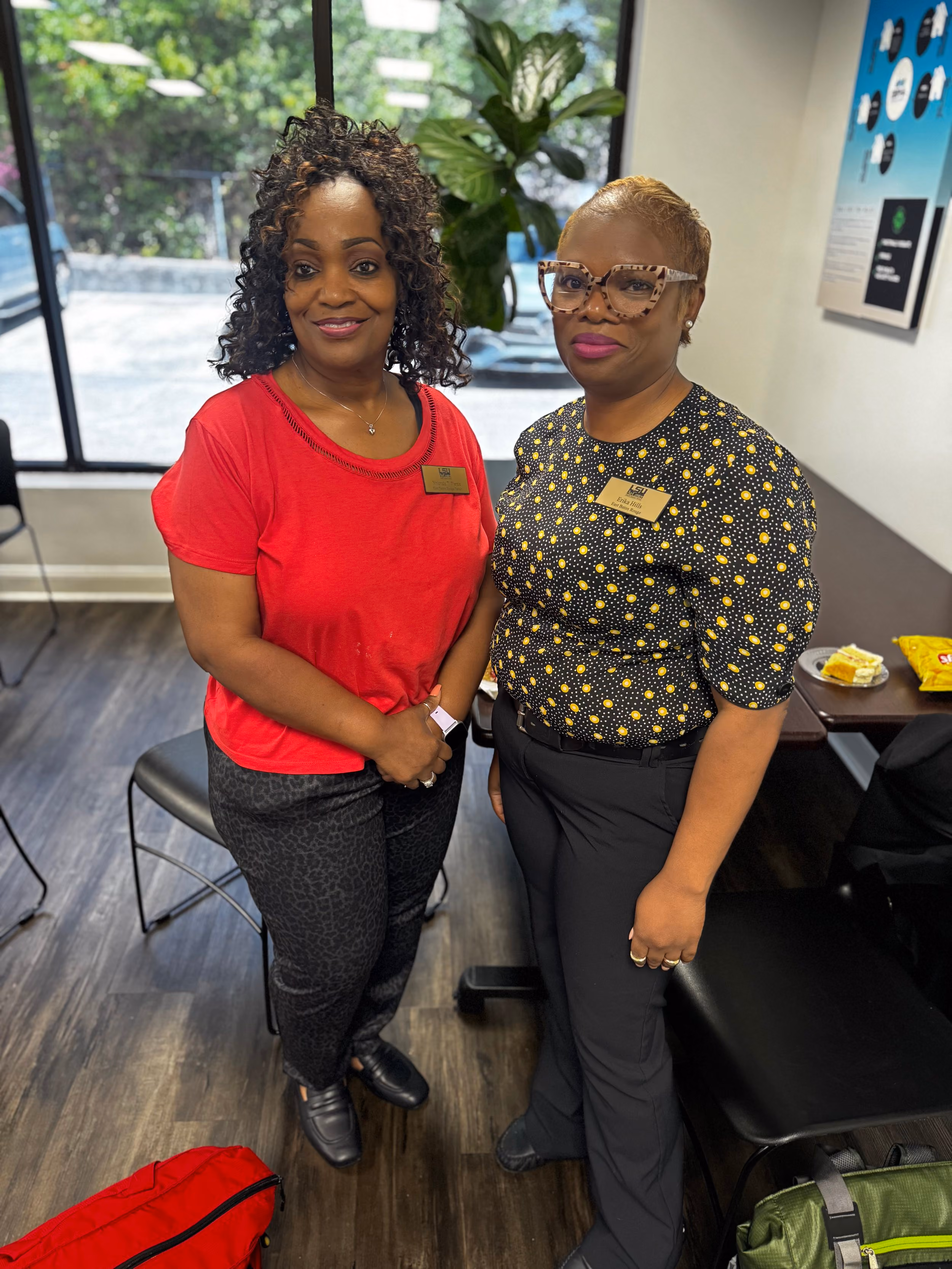 Two women standing in an office space, smiling at the camera. The woman on the left wears a red shirt and black patterned pants, and the woman on the right wears a black and yellow polka-dotted blouse with black pants and large glasses. They both have name badges. The background shows a window, chairs, a plant, and a table with snacks.