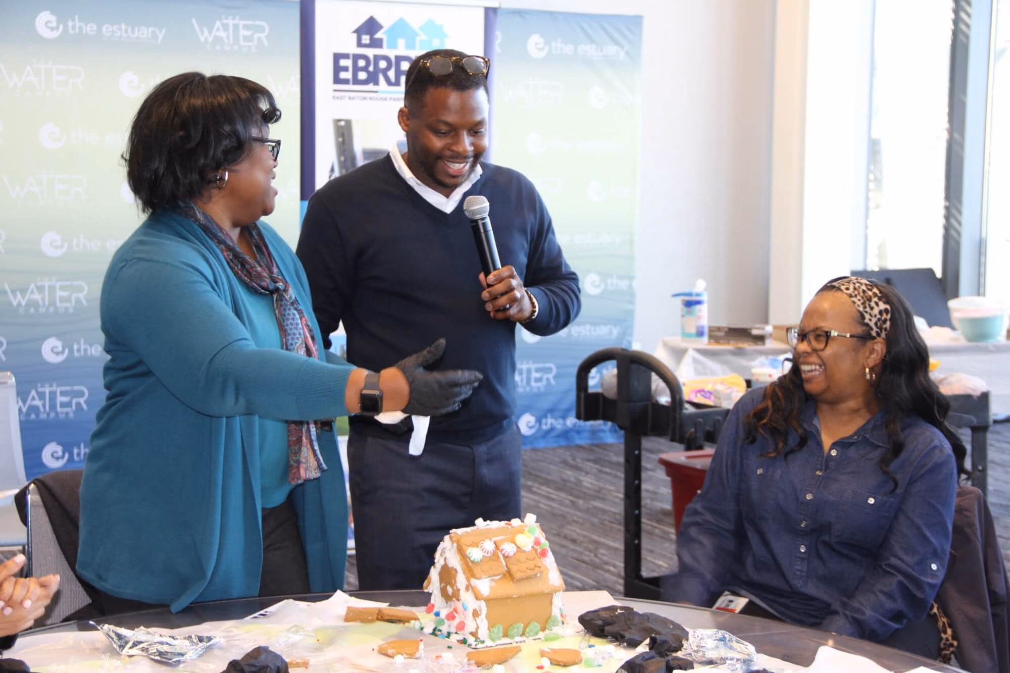 Three people smiling and interacting around a table with a decorated gingerbread house; one man holds a microphone, a woman in a teal jacket gestures towards the house, and another woman in a navy shirt and leopard headband sits laughing.