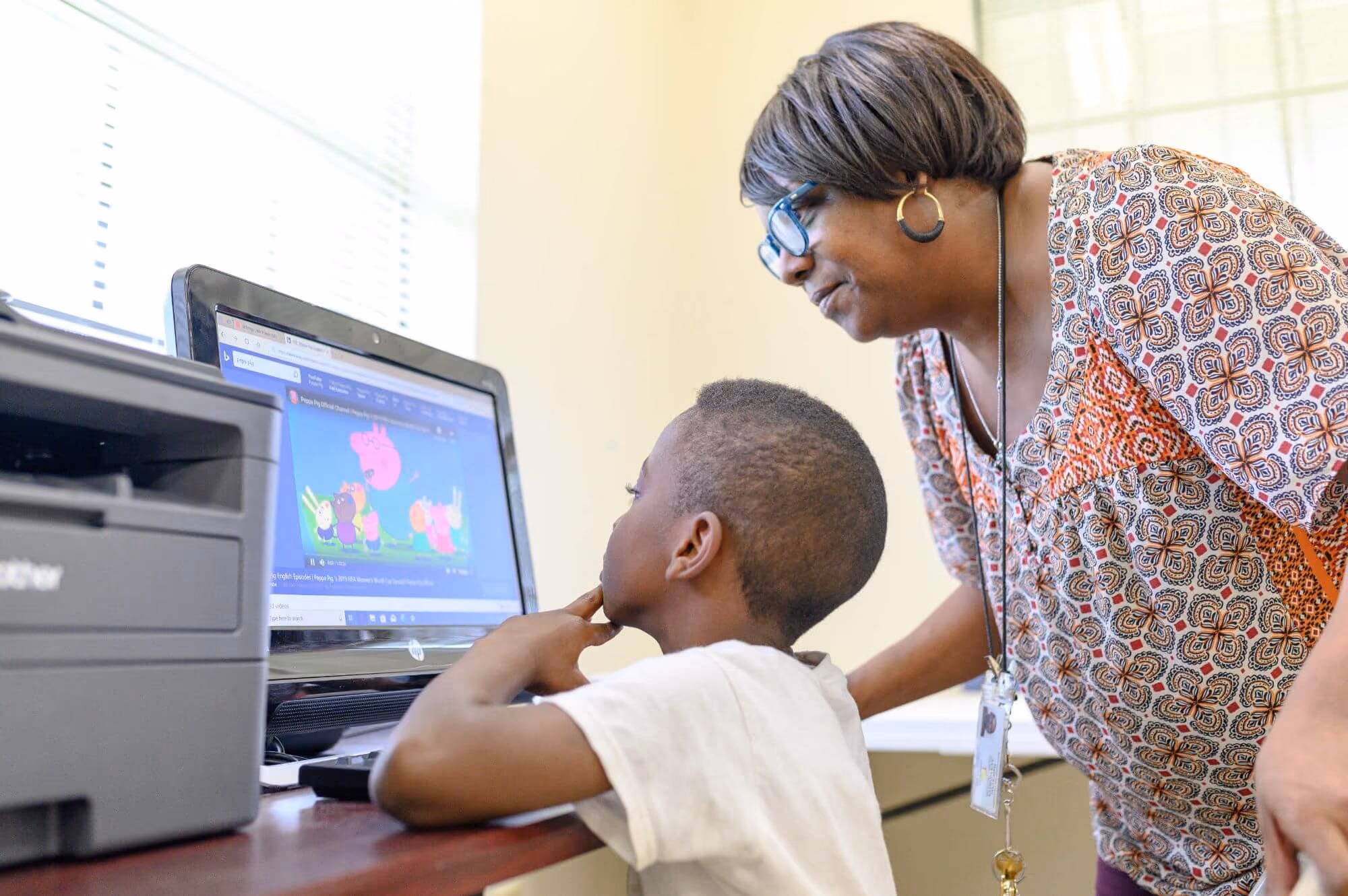 A young boy sitting at a desk in front of a desktop computer watching an educational cartoon while an adult woman, possibly a teacher or guardian, leans over him observing the screen.