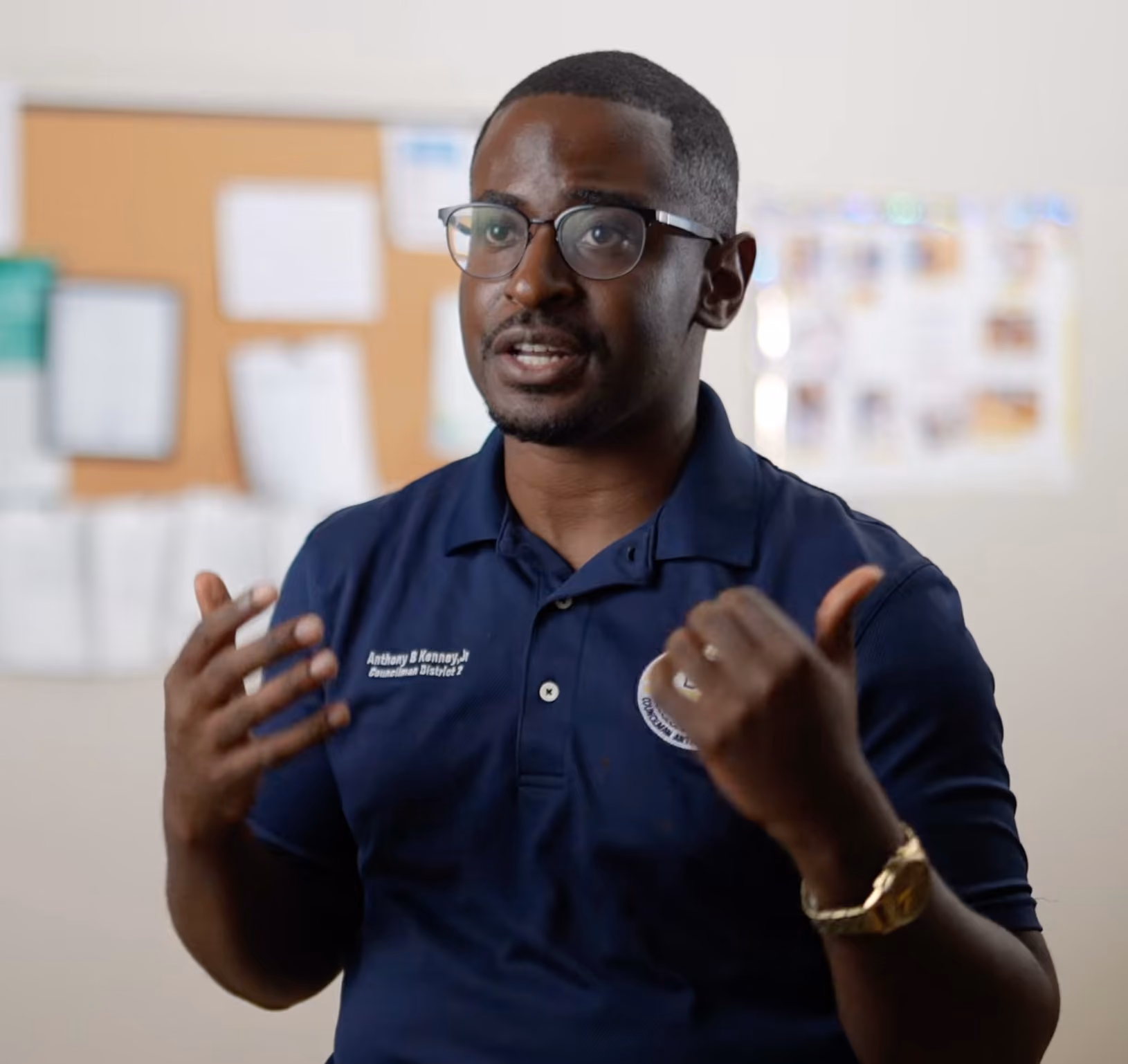 Man wearing glasses and a navy blue polo shirt with a name and title, gesturing with his hands while speaking in an indoor setting with a blurred bulletin board in the background.