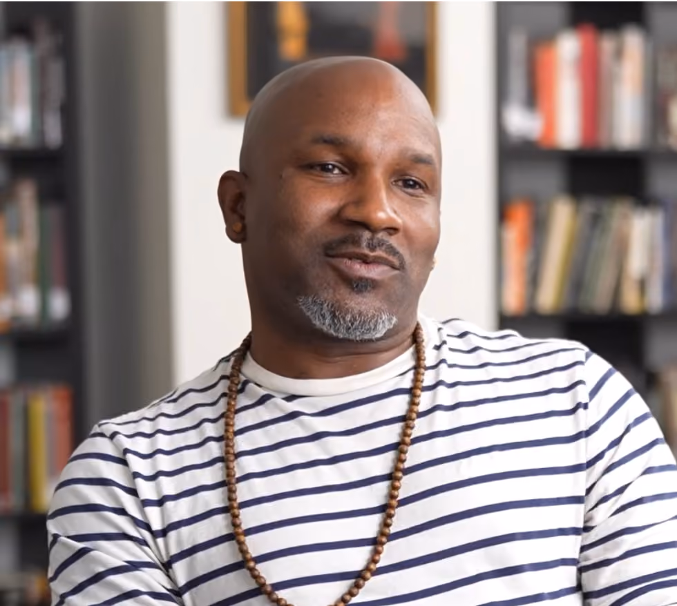 A bald man with a gray goatee wearing a white and navy striped shirt and a wooden bead necklace, sitting indoors with blurred bookshelves in the background.