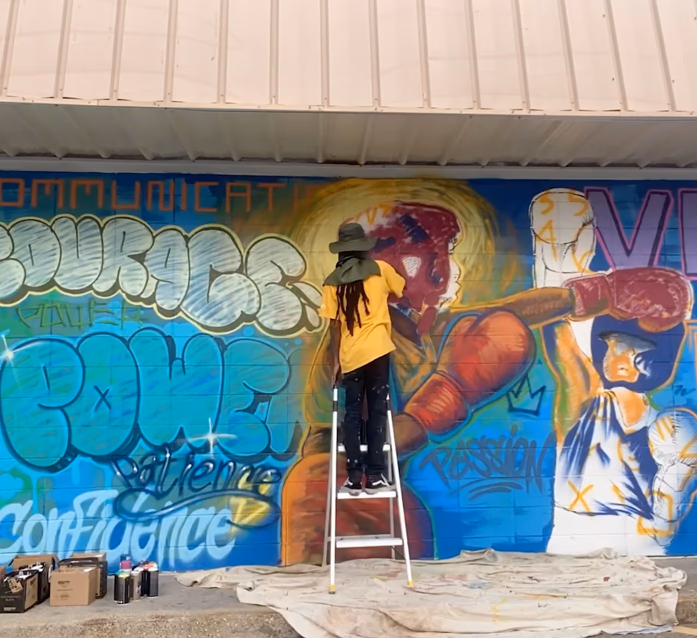 A person with dreadlocks wearing a wide-brimmed hat and a yellow shirt stands on a ladder painting a colorful graffiti mural of two boxers wearing gloves on a wall filled with words like 'courage,' 'power,' and 'confidence.'