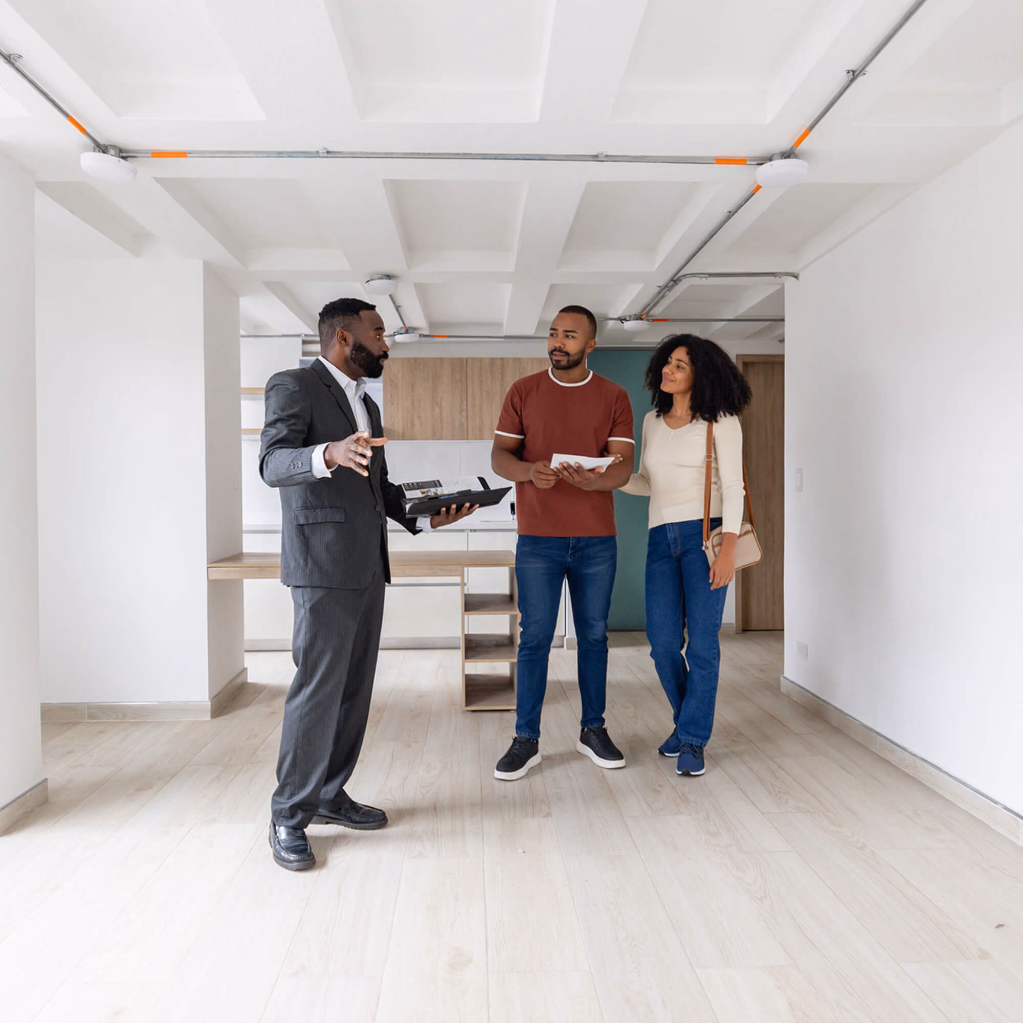 A real estate agent in a suit showing an apartment to a couple standing in an empty room with wooden flooring and white walls. The man holds a tablet, and the woman stands beside him carrying a beige shoulder bag.