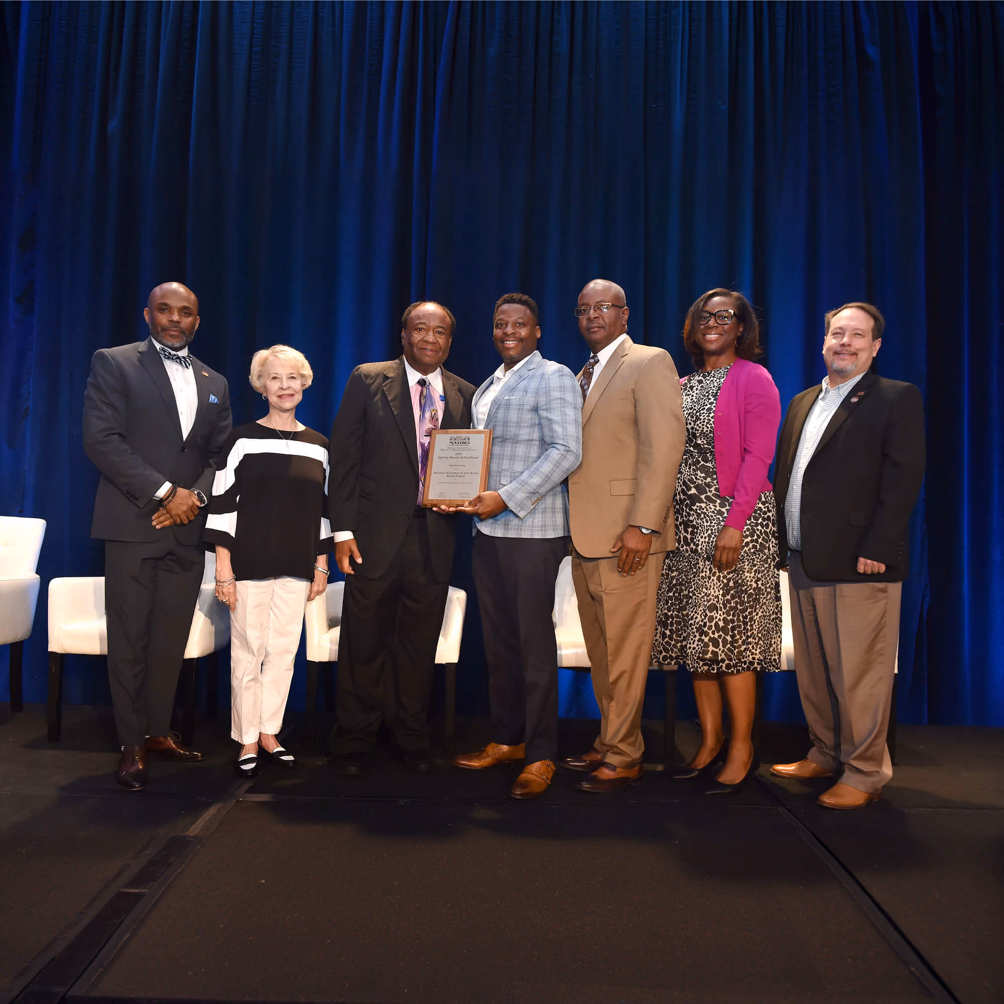 Seven people standing on a stage with a dark blue curtain backdrop, five men and two women dressed in business attire, with the fourth person from the left holding a framed award plaque and smiling.