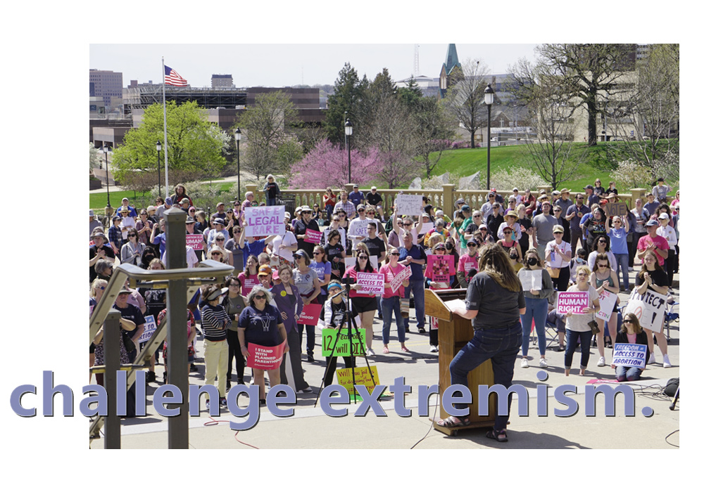A large group of people gathered at the Iowa State Capitol to rally in favor of reproductive rights for all, many of whom are holding signs. They are listening to a woman at the podium who is speaking about how we can all use our voices to make change.
