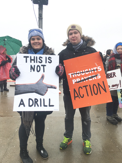 Two protesters look at the camera while holding their signs that read "This is not a drill" with an image of a hand pointed like a gun at the other sign, which reads, "Thoughts Prayers ACTION" -- both Thoughts and Prayers are crossed out.