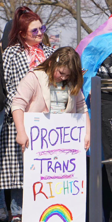 A young person looks down at a sign they've made that says "Protect Trans Rights!" with a rainbow at the bottom.