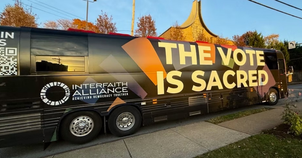 A large black coach bus with "The Vote is Sacred" printed in large yellow letters on the side. In a smaller font it says "Interfaith Alliance: Achieving Democracy Together."