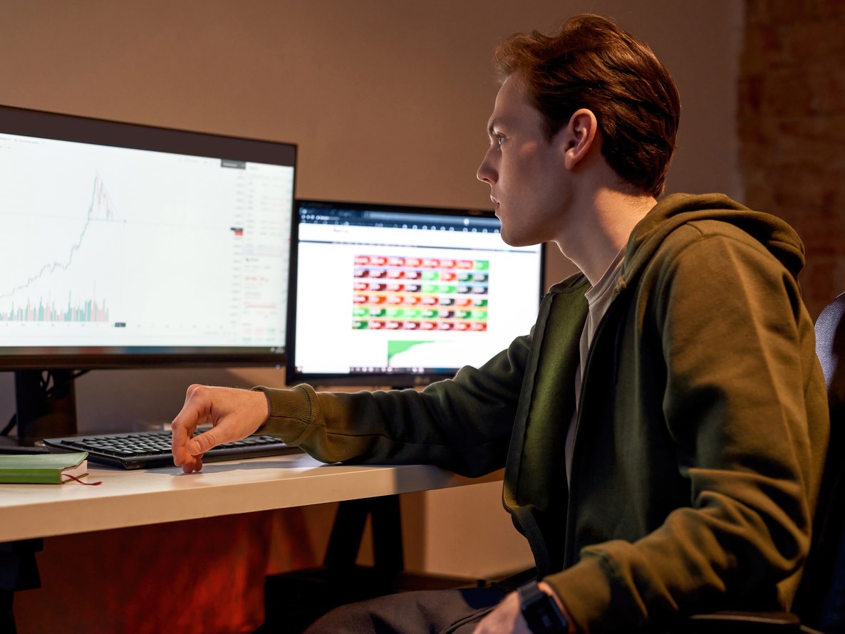 Trader joven opera desde su escritorio con dos monitores, uno con un gráfico de velas y otro con la plataforma abierta