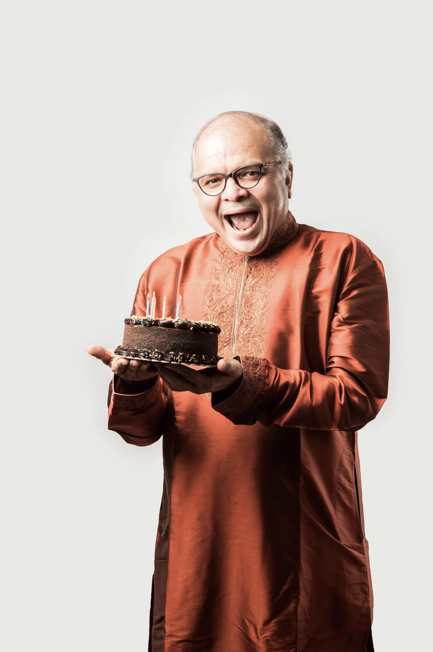 Smiling older man in traditional clothing holding a chocolate birthday cake with candles.