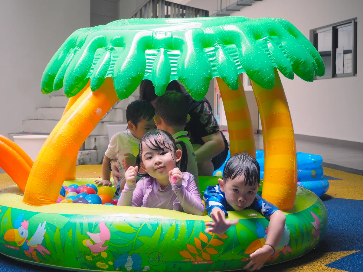 Children playing in an inflatable pool with a green palm tree canopy indoors.