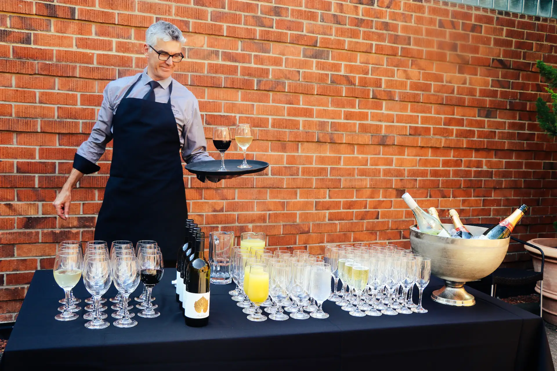 Man in black apron and glasses standing behind a table with rows of clear empty wine glasses, filled wine glasses, bottles of wine, and pitchers of orange juice and water in front of a brick wall, holding a tray with one glass of white wine and one glass of red wine.