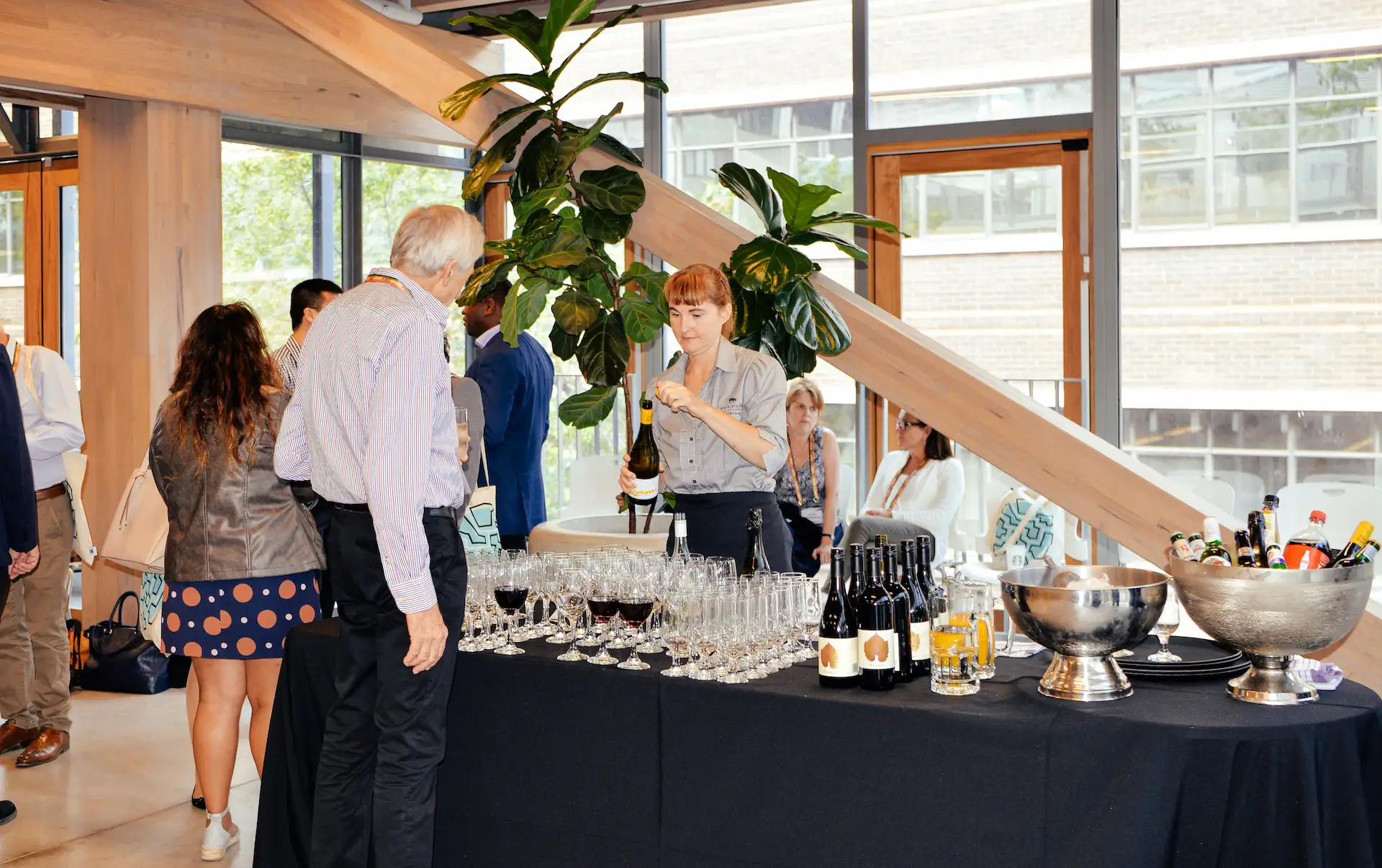 A woman behind a table with a black tablecloth is opening a bottle of white wine while a man in a striped shirt and black pants watches; many wine glasses, bottles of red wine, and pitchers of orange juice are arranged on the table, with large silver bowls filled with bottles and beverages to the right, set in a modern indoor space with wooden beams and large windows.