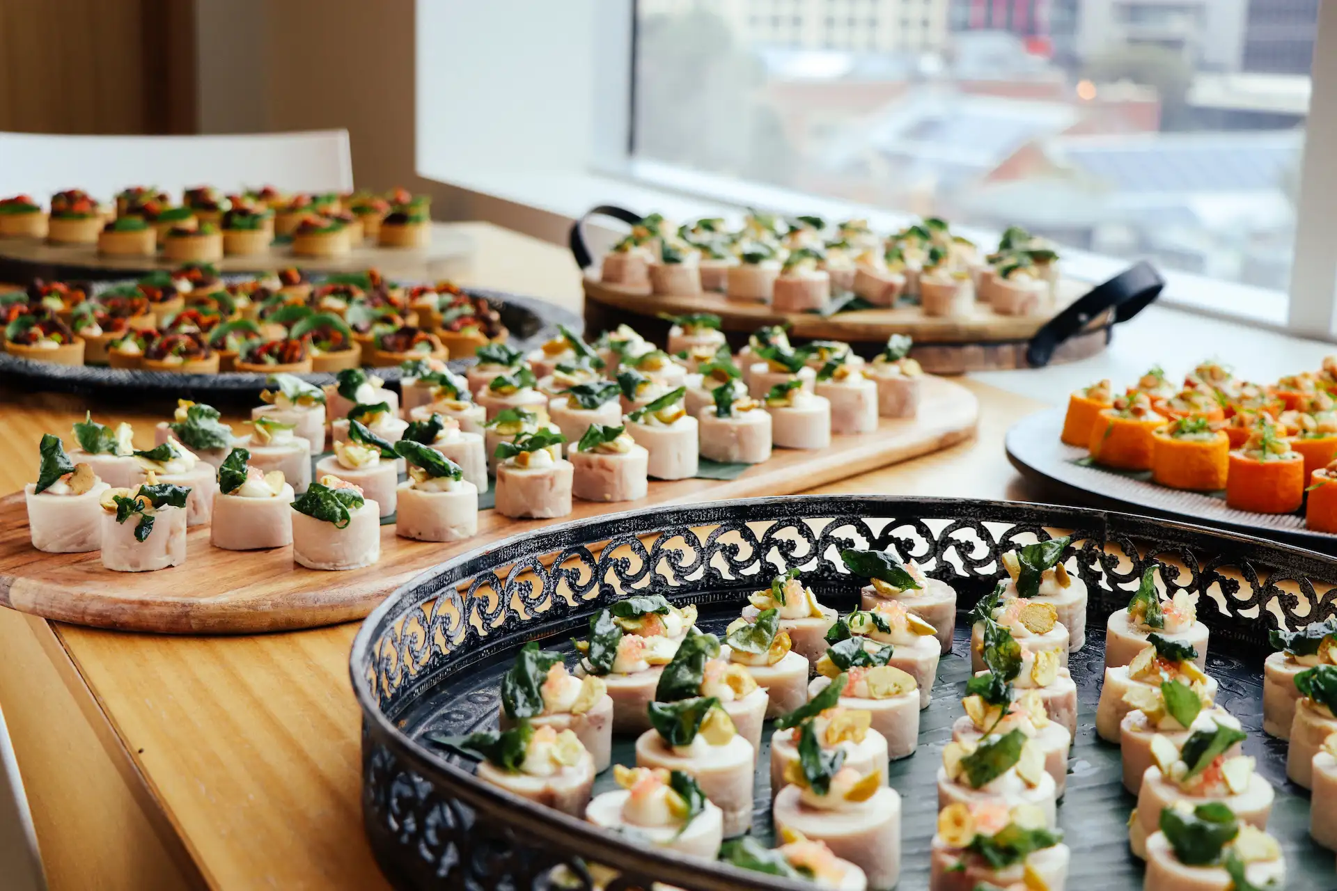 Multiple platters of assorted elegant hors d'oeuvres arranged on a wooden table by a large window with an urban view, including small rolled appetizers garnished with leafy greens and seeds, and brightly colored bite-sized pieces.