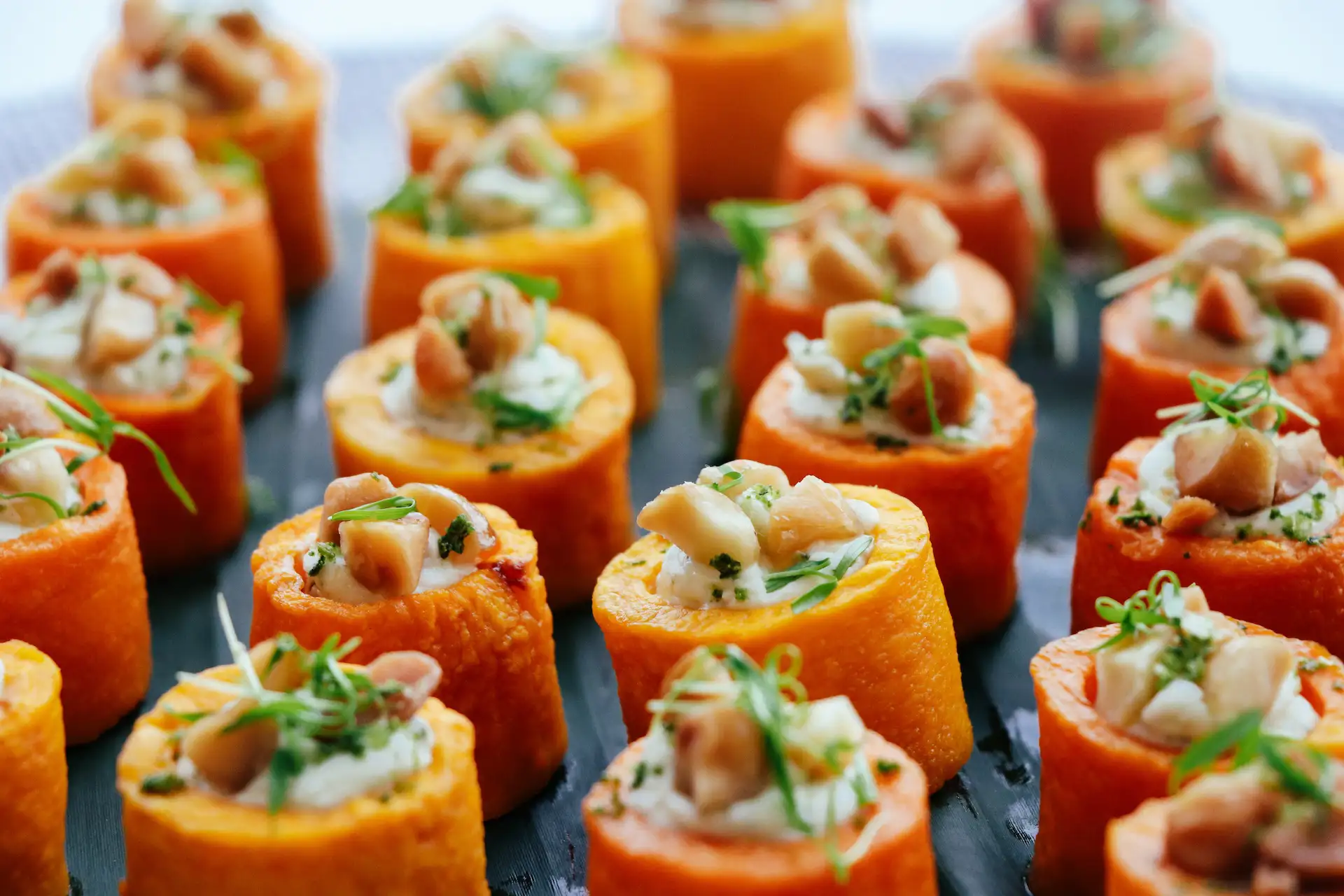 Orange-colored bite-sized appetizers arranged on a dark surface, each topped with white cream, chopped nuts, and green herbs.