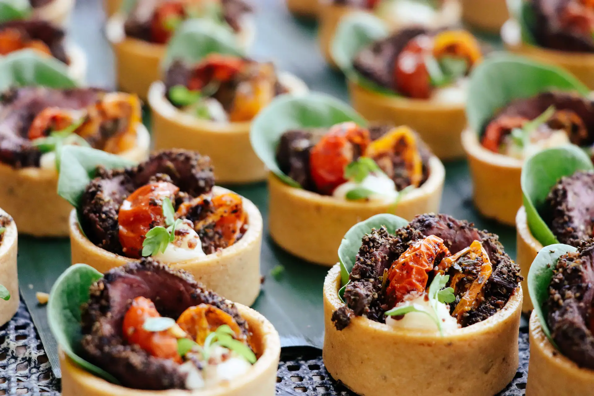 Close-up of multiple savory tartlets filled with slices of seasoned meat, roasted cherry tomatoes, leafy greens, and garnished with fresh herbs, arranged on a dark surface.