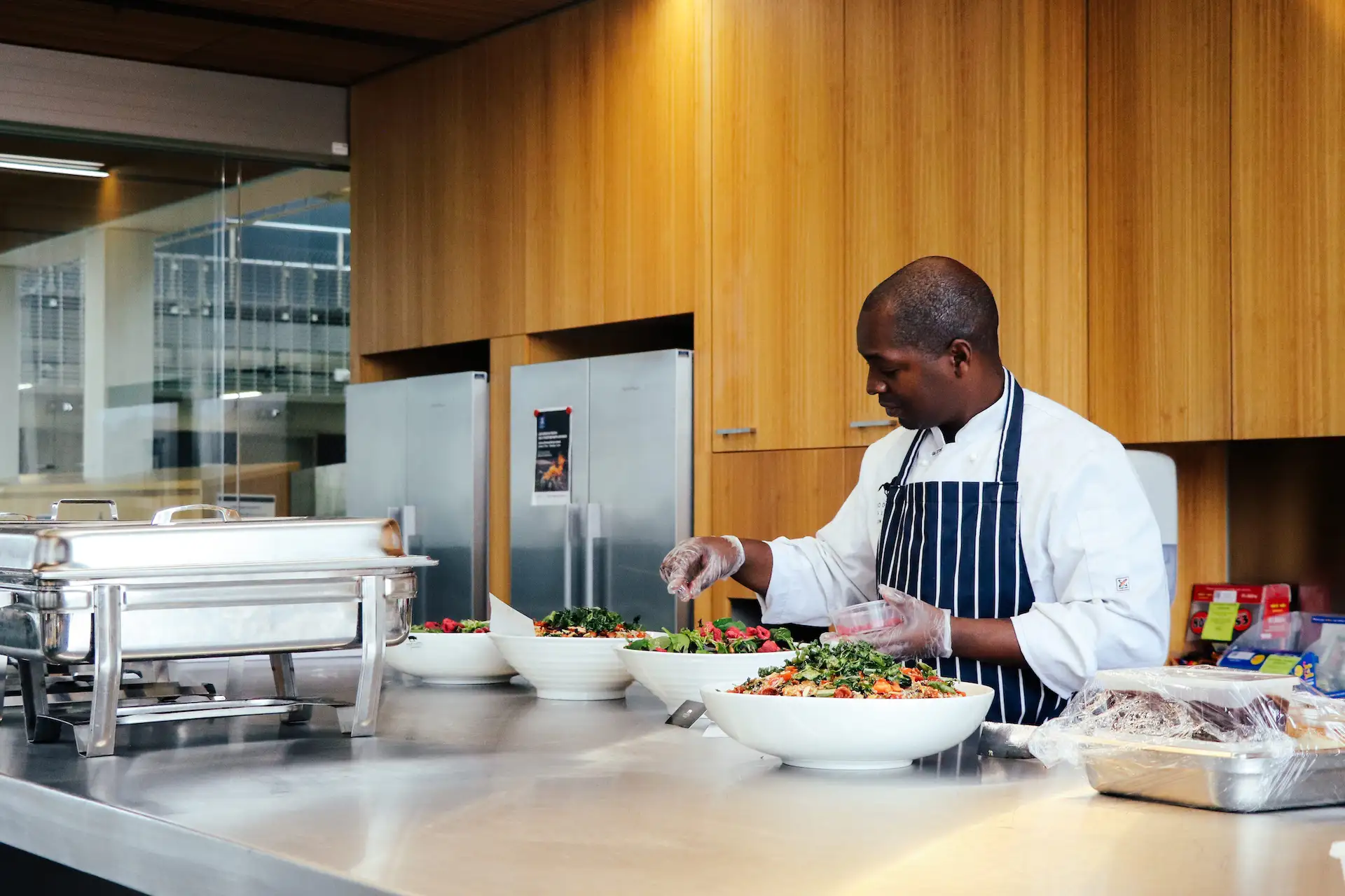 Chef wearing a white coat and striped apron preparing multiple large bowls of fresh salad on a stainless steel kitchen counter in a modern kitchen with wooden cabinets and stainless steel refrigerators.