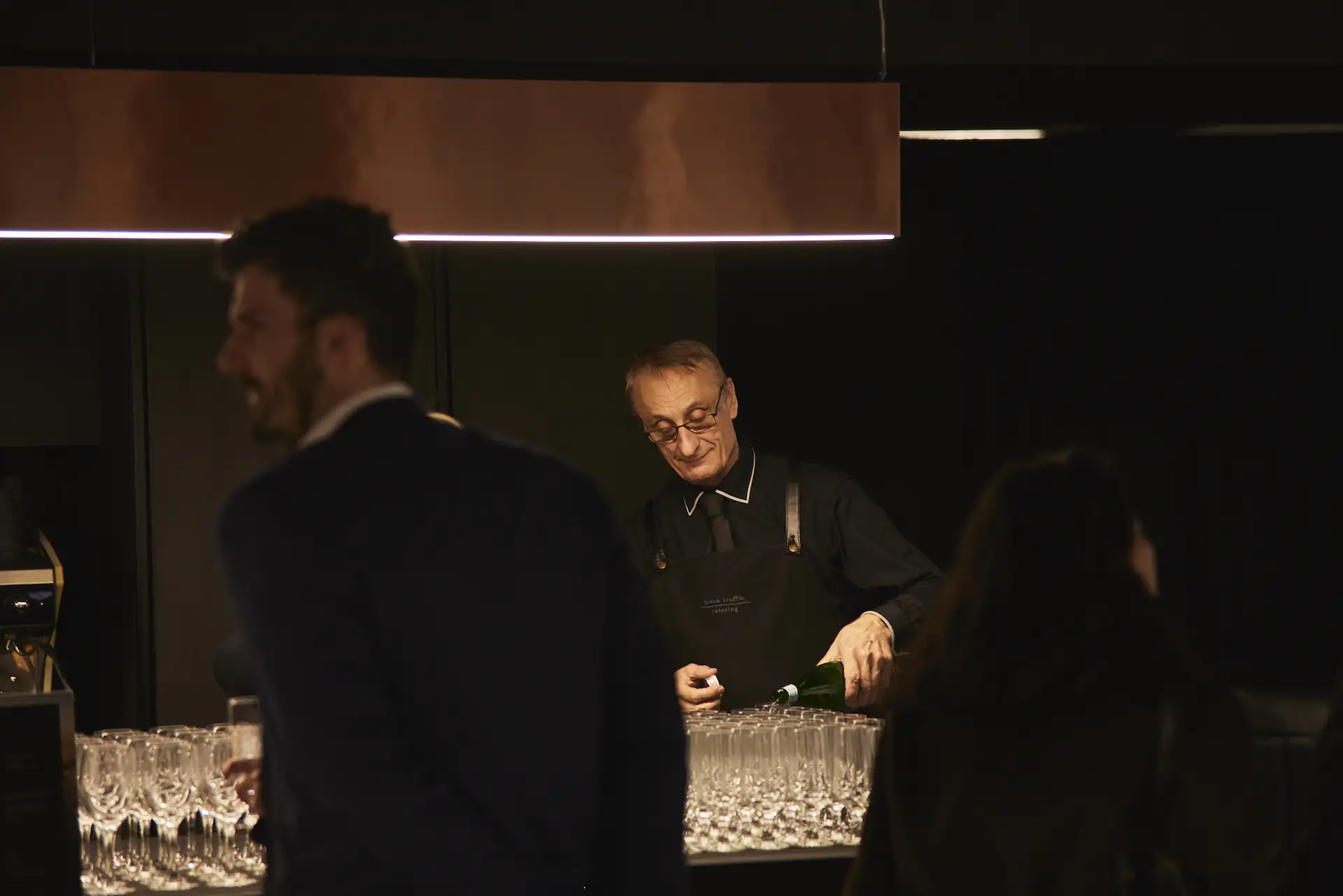 Bartender wearing glasses and a black apron pouring a drink into empty glasses arranged on the bar counter with two blurred patrons in the foreground.