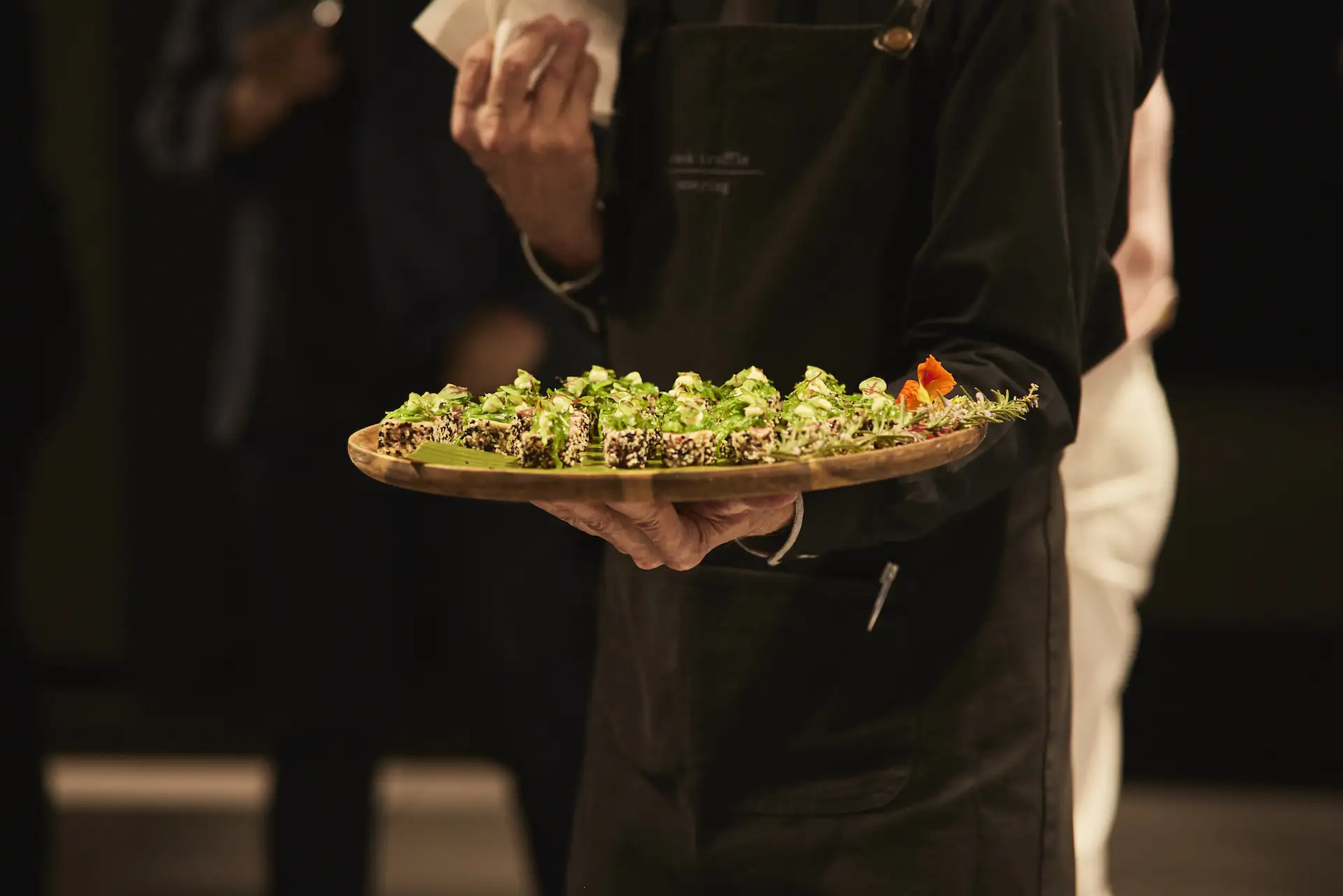 Waiter wearing black apron holding a wooden tray with neatly arranged bite-sized appetizers garnished with green herbs and a small orange flower on a dark background.