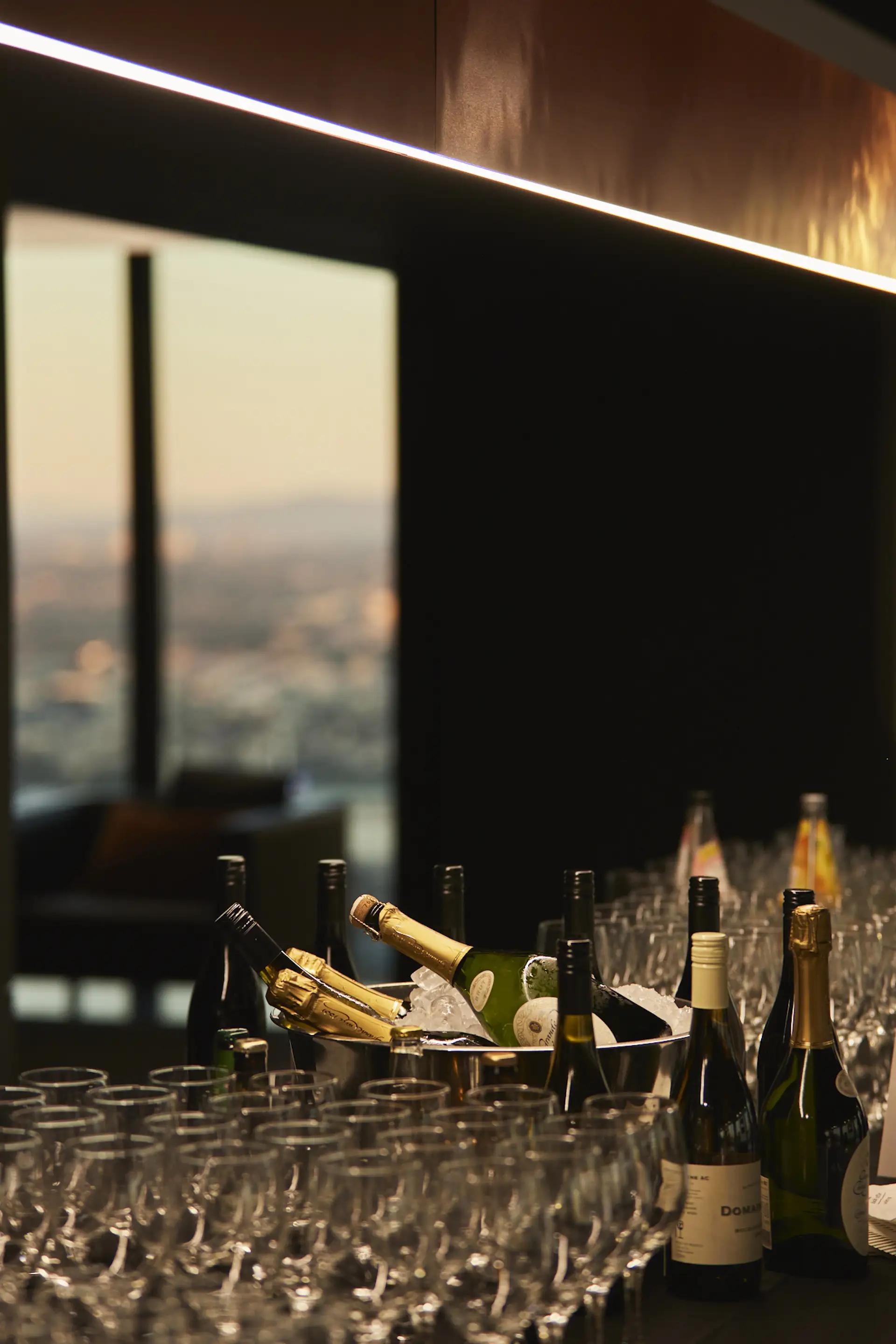 Table with multiple wine glasses and bottles, including champagne bottles chilled in an ice bucket, set in a dimly lit room with a window showing an out-of-focus cityscape in the background.