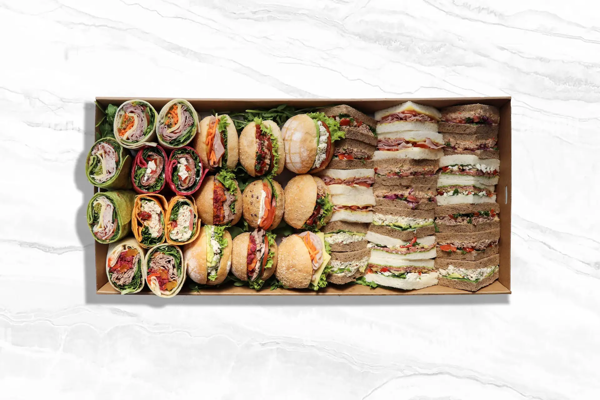 Assorted lunch platter with an array of wraps, mini sandwich buns with various fillings, and stacked finger sandwiches on white and brown bread, all arranged neatly in a rectangular box on a white marble surface.