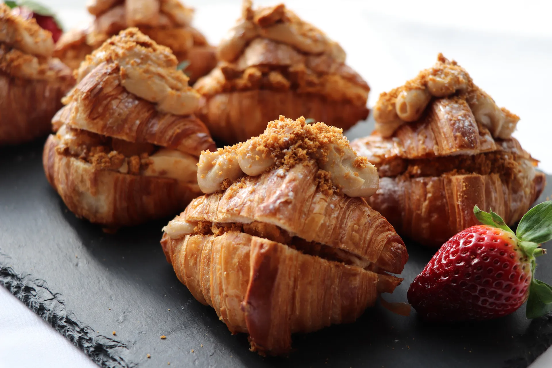 Several croissants filled and topped with a creamy spread and sprinkled with crumbs, arranged on a dark slate platter with a fresh strawberry on the side.
