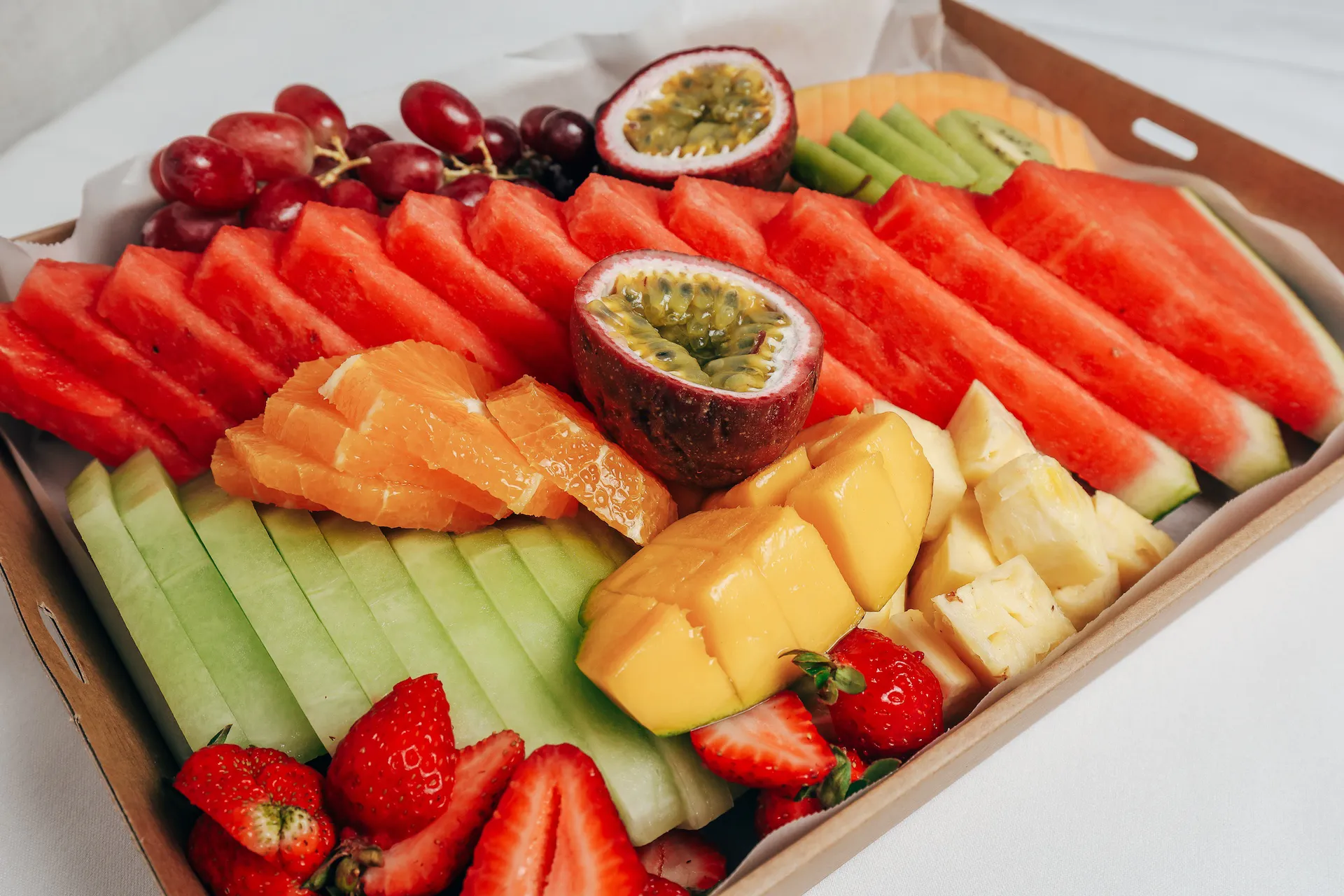Tray of assorted fresh fruits including sliced watermelon, passion fruit halves, orange slices, honeydew melon slices, mango chunks, pineapple pieces, strawberries, grapes, and kiwi slices.