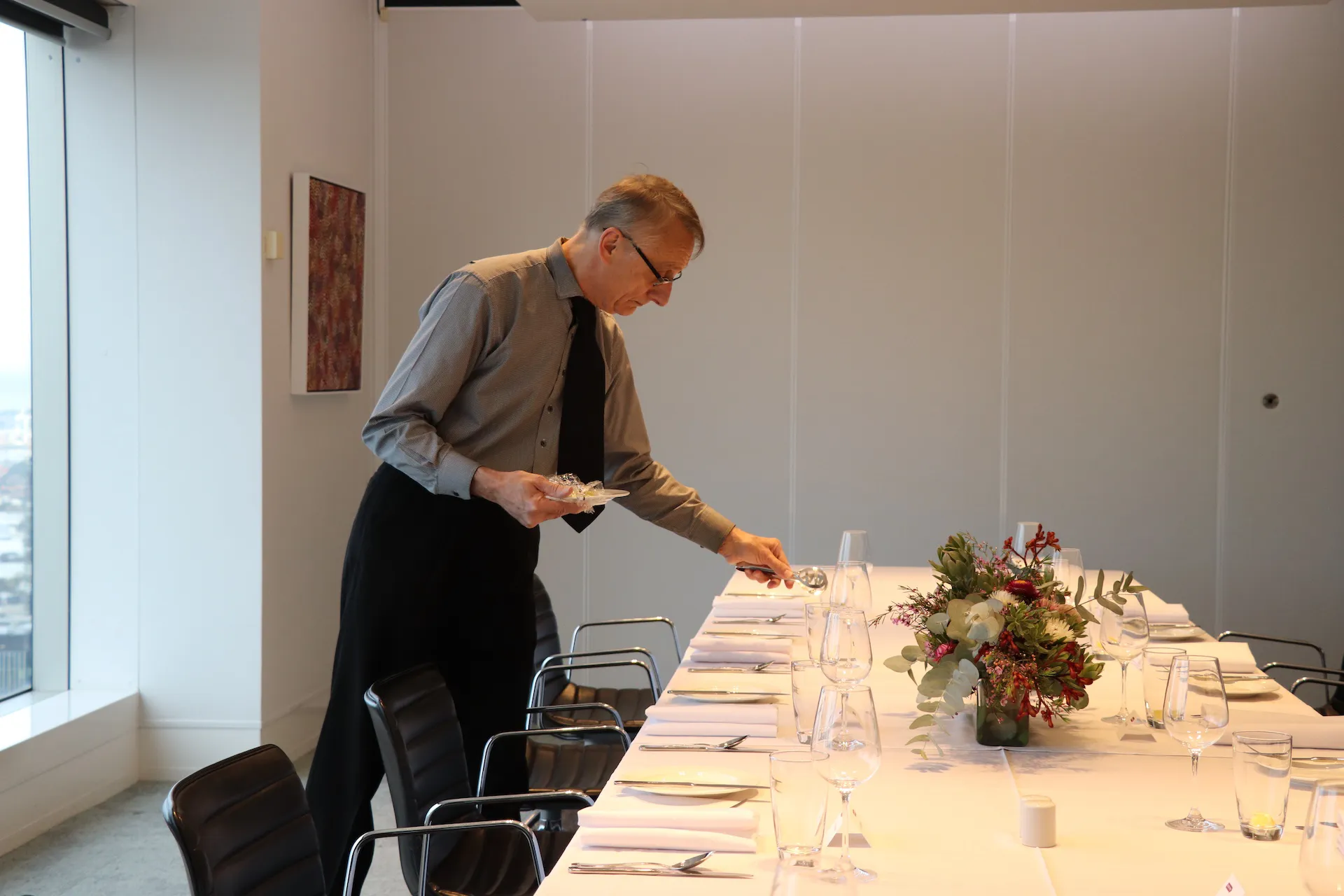 Man wearing glasses, gray shirt, black tie, and apron sets a long, white-clothed dining table in a modern meeting room with black chairs and a bouquet centerpiece.