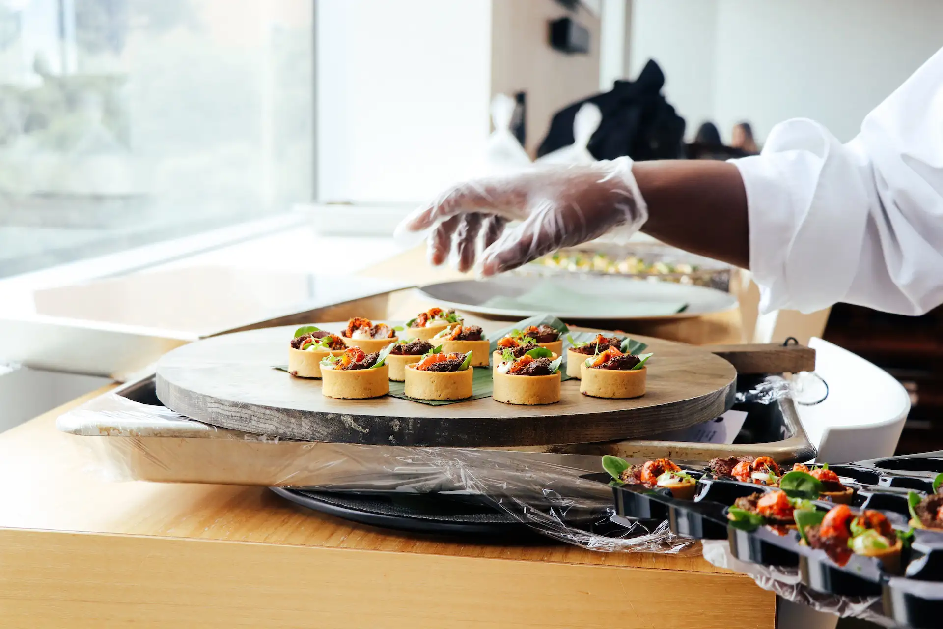 Close-up of a catering staff member wearing a white shirt and glove arranging small savory tart appetizers on a round wooden serving board at a well-lit event setting.